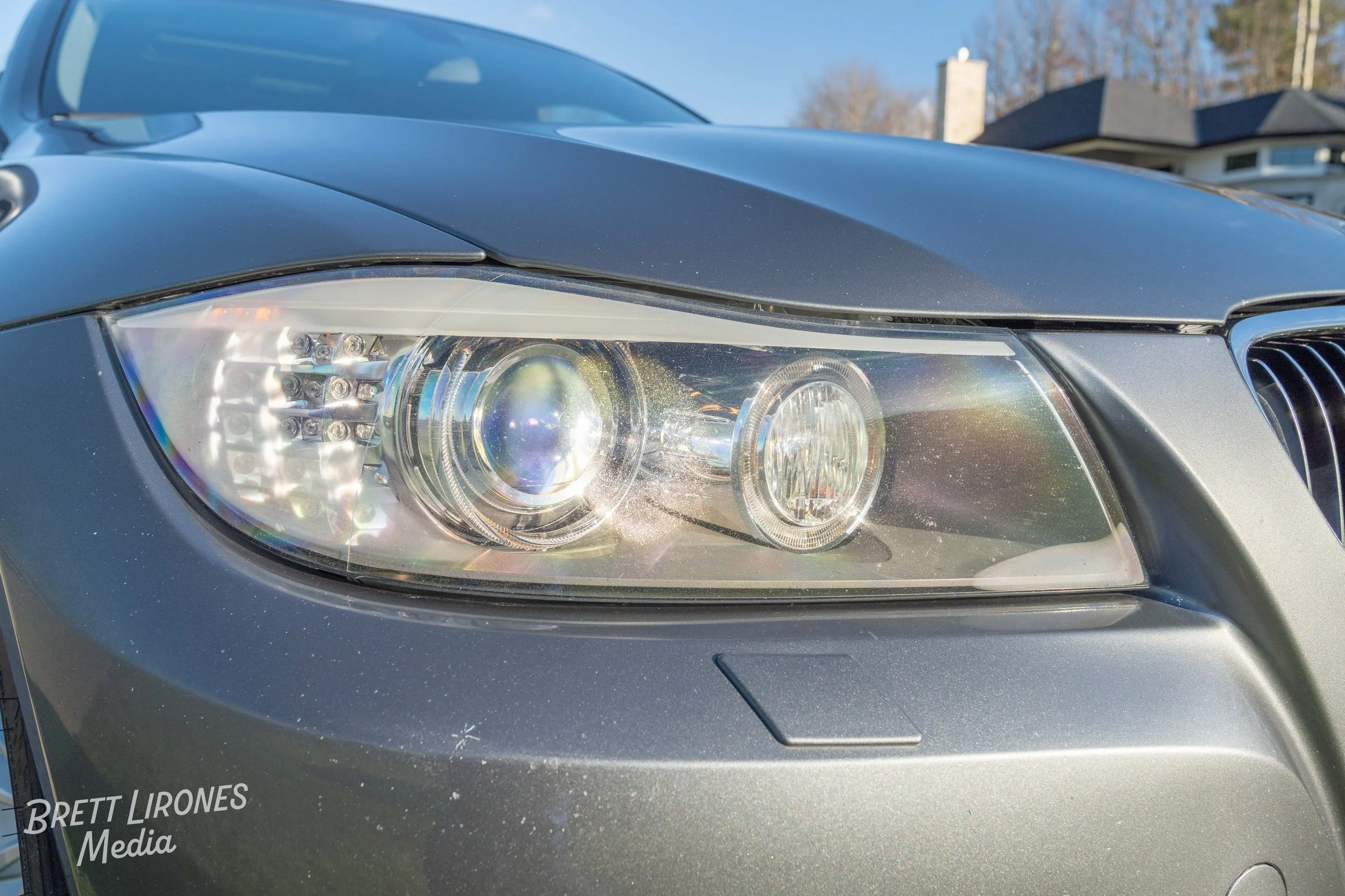 Close-up of a car's front headlight on a silver vehicle, with a house and trees in the background.