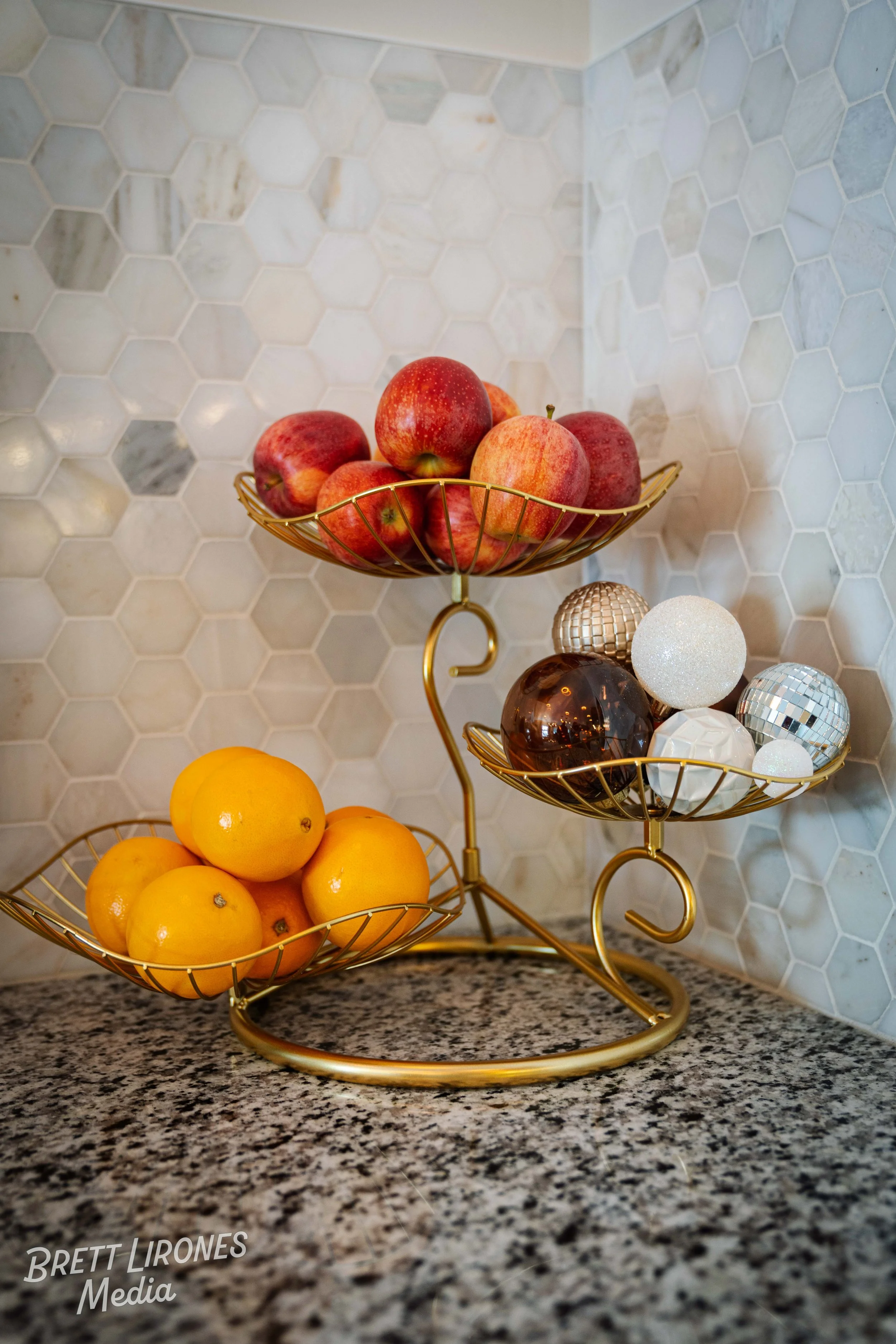 A gold, three-tiered fruit and ornament holder with red apples on the top tier, yellow lemons on the bottom tier, and various decorative spheres on the middle tier, set against a white hexagonal tile backsplash on a granite countertop.