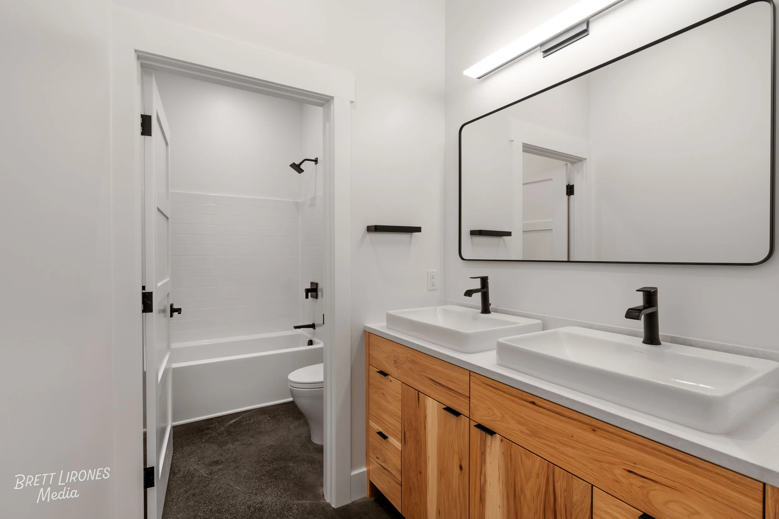Modern bathroom with double vanity sinks, wooden cabinetry, large mirror, black fixtures, and a separate shower area with a bathtub and black showerhead.
