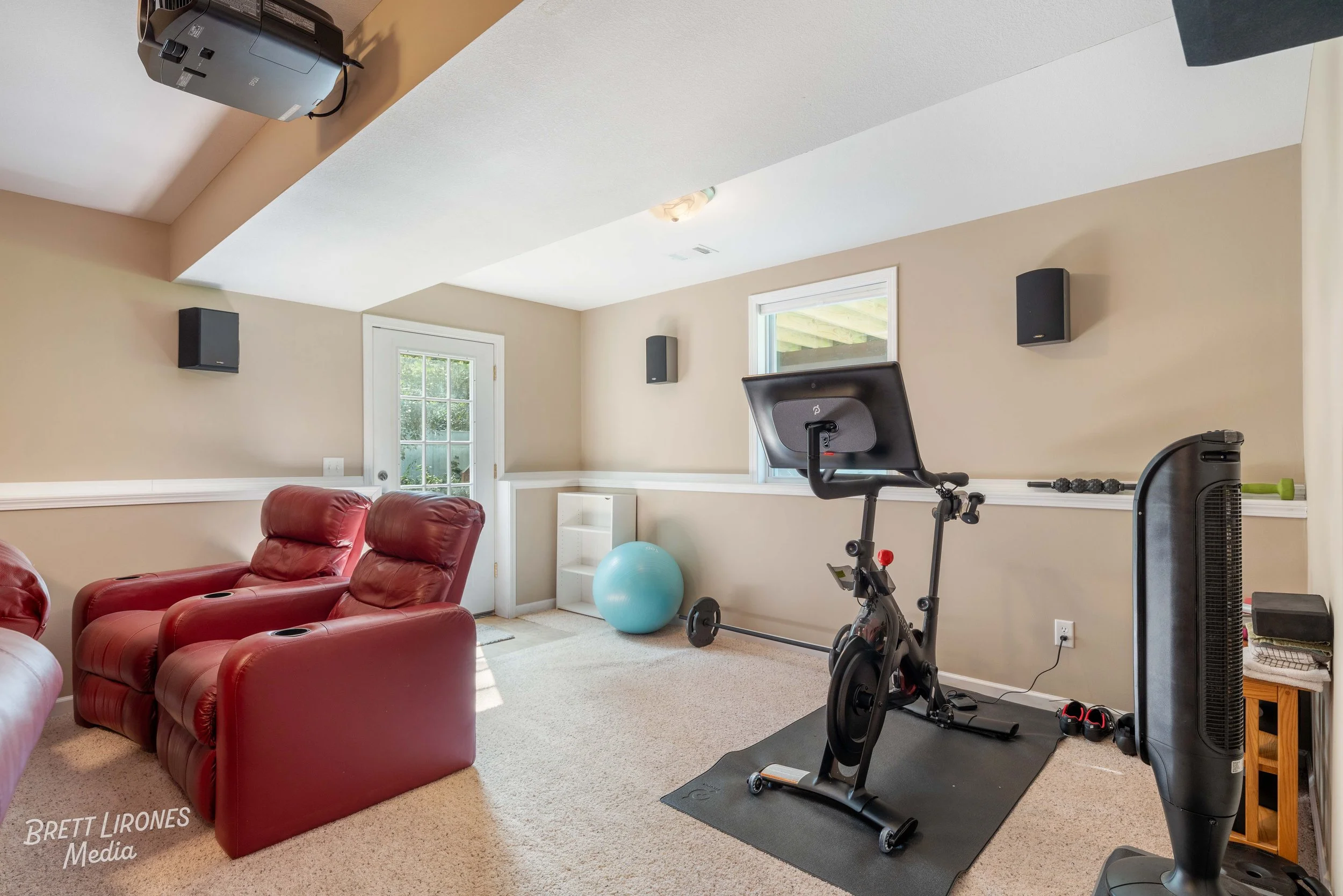 A basement room with beige walls and carpeted floor, containing a red recliner sofa, a stationary exercise bike, a blue exercise ball, a white shelving unit, a window and a glass door, and black wall-mounted speakers.
