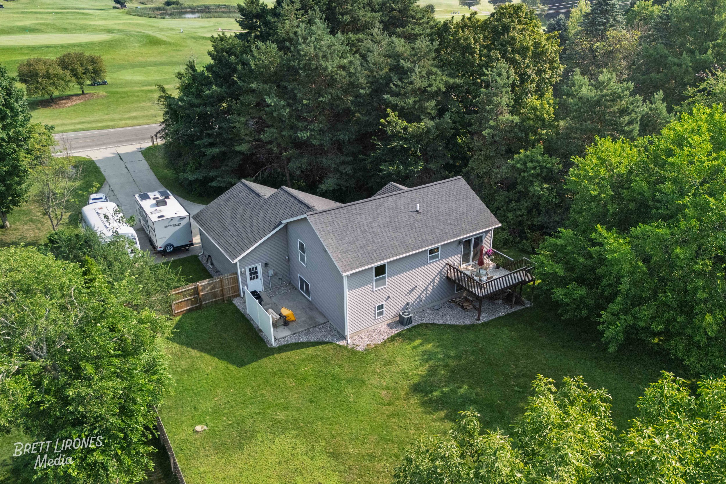 An aerial view of a house with a backyard deck, surrounded by trees and a grassy yard, with a gravel driveway parking two vehicles nearby.
