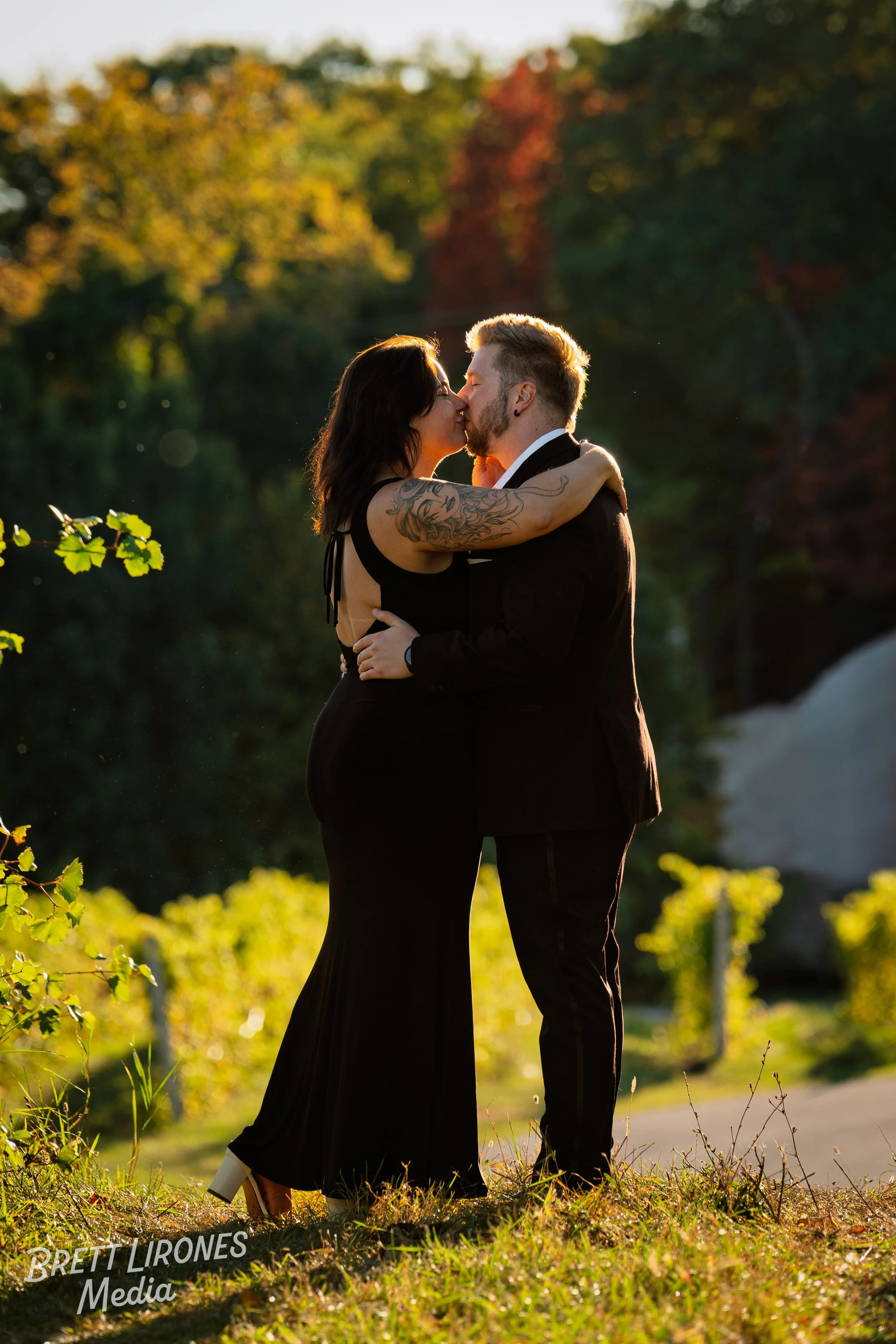 A couple dressed in formal attire embracing and about to kiss outdoors during sunset with a background of trees with fall foliage.