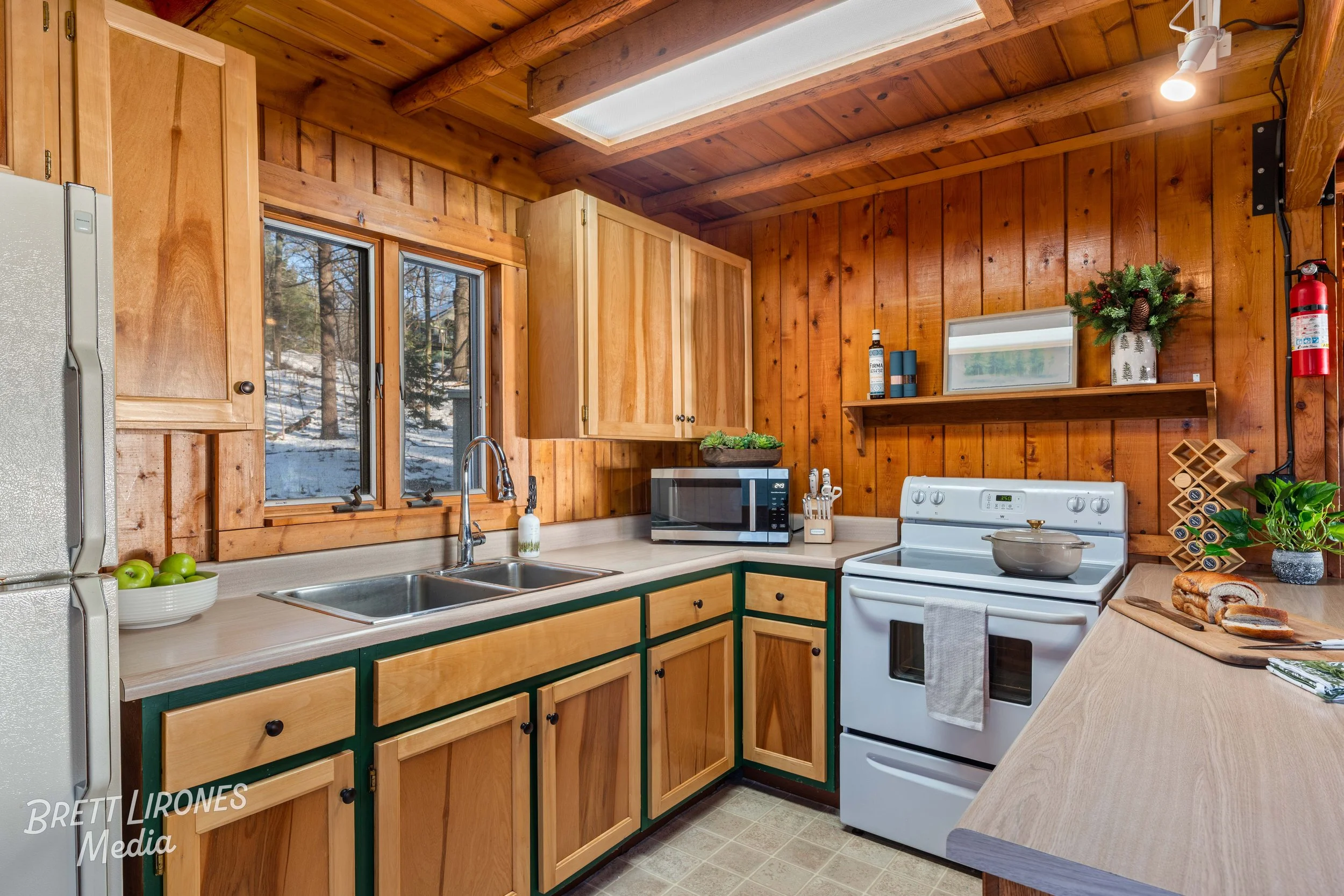 Wood-paneled kitchen with window overlooking outdoors, white stove, microwave, kitchen counter with bread, green apples, and potted plants, with holiday decorations, cabinets, and a skylight ceiling.