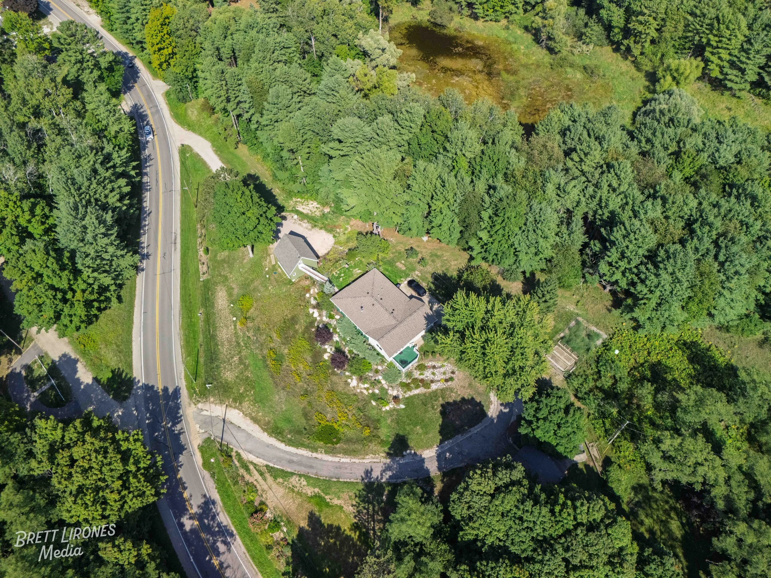 An aerial view of a small house surrounded by lush green trees, a driveway, and a garden on a hillside near a winding road.