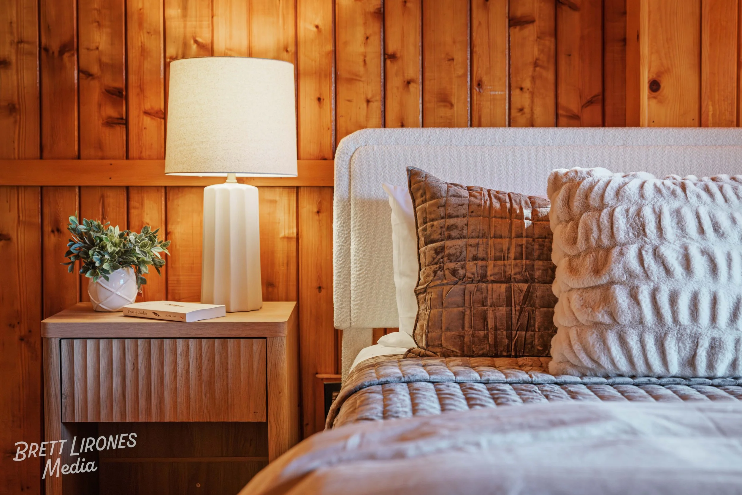 A cozy bedroom with a bed featuring brown and beige textured pillows, a wooden nightstand with a potted plant, books, and a white lamp, against a wooden-paneled wall.