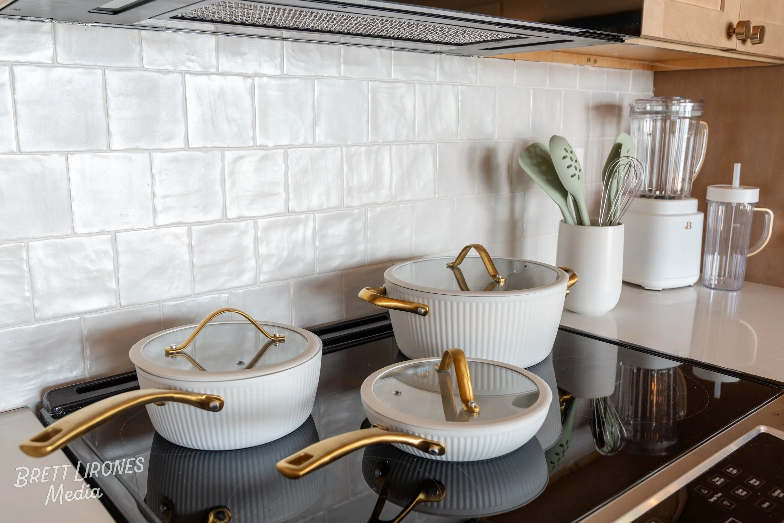 Kitchen stovetop with three white ceramic cookware with gold handles and glass lids, a utensil holder with green utensils, a blender, and a glass jar on a white countertop against a white tiled backsplash.