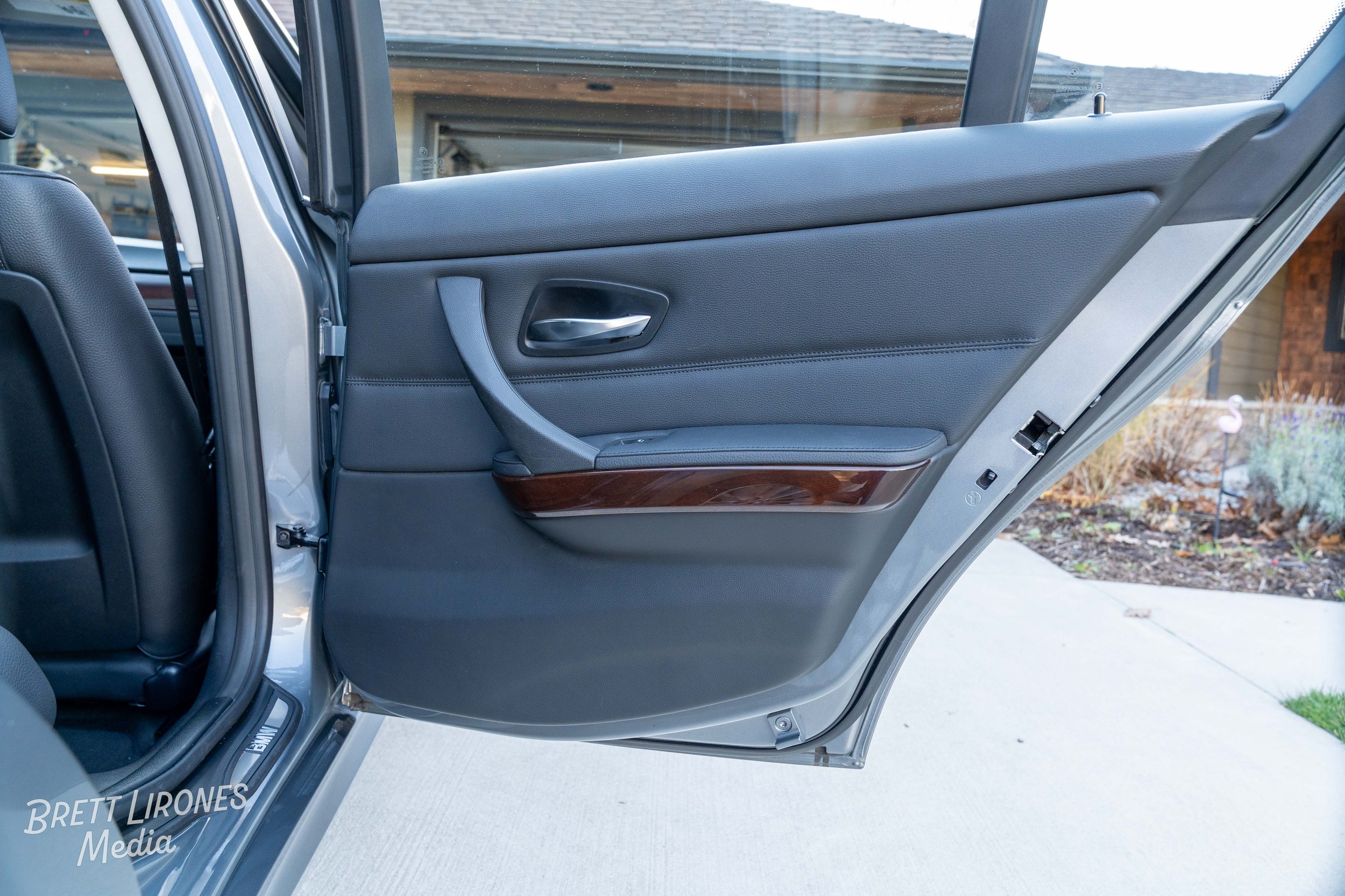 The inside of a car door with a gray interior, window, and a wood trim detail, parked in a driveway with a house and garden in the background.