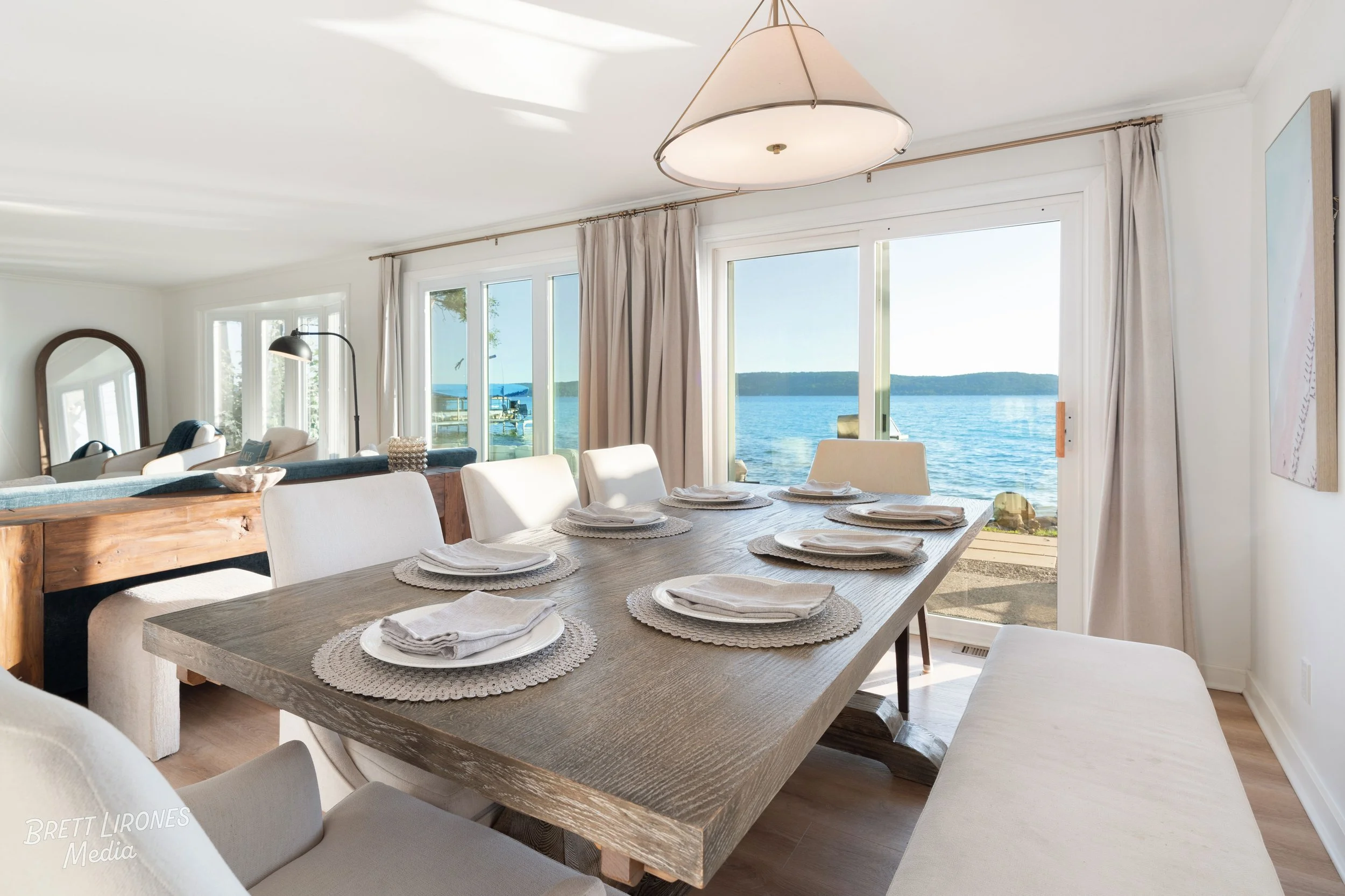 Dining room with large wooden table set with white plates, napkins, and woven placemats, overlooking a lake through glass sliding doors, with light-colored chairs, curtains, and a pendant ceiling light.