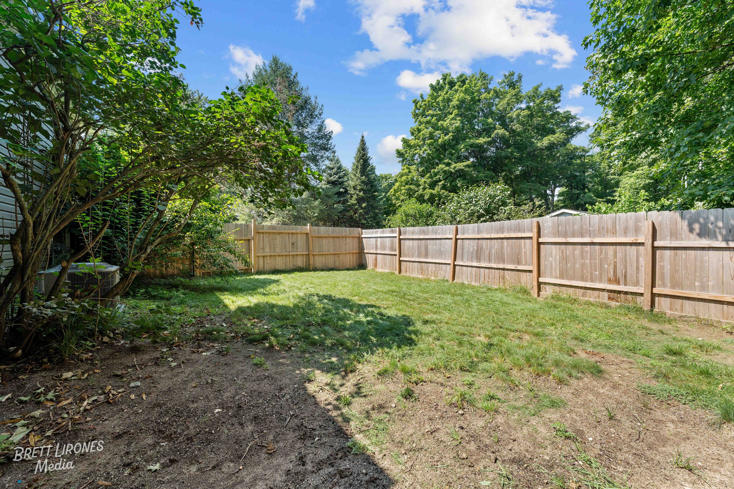 A backyard with a grassy lawn, a wooden fence, and green trees against a partly cloudy sky.