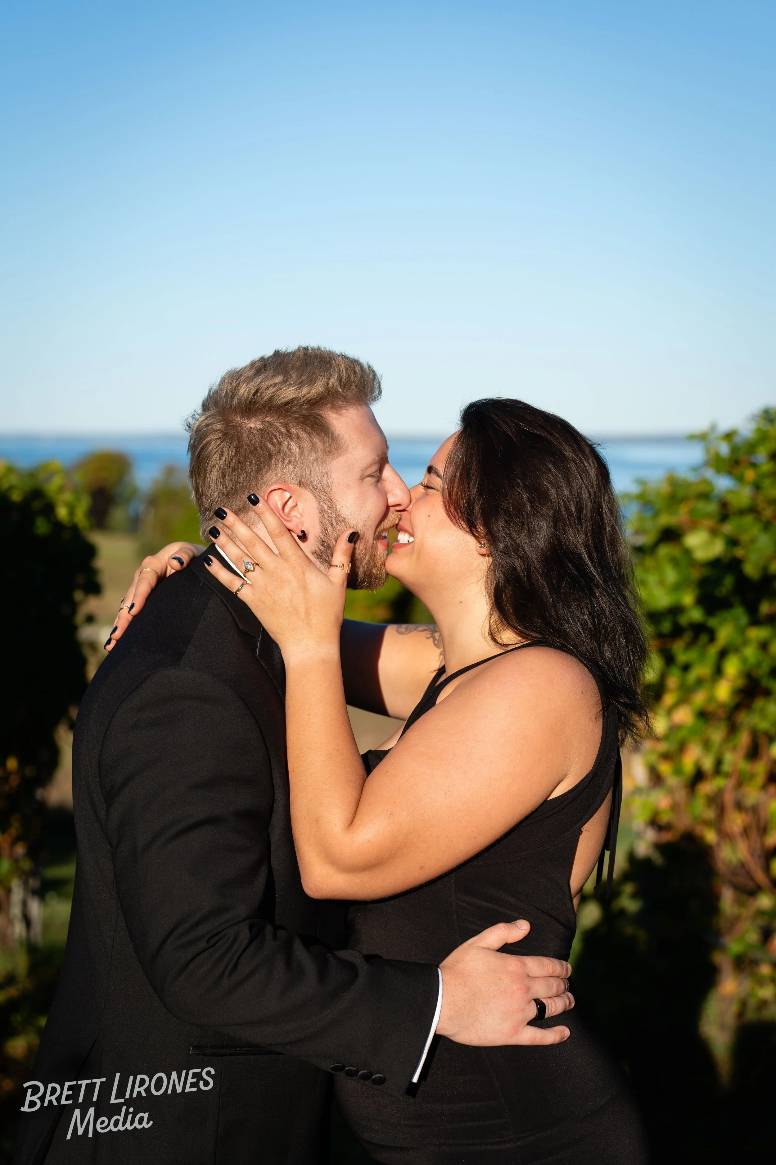 A couple dressed in black sharing an intimate kiss outdoors with a blue sky and greenery in the background.