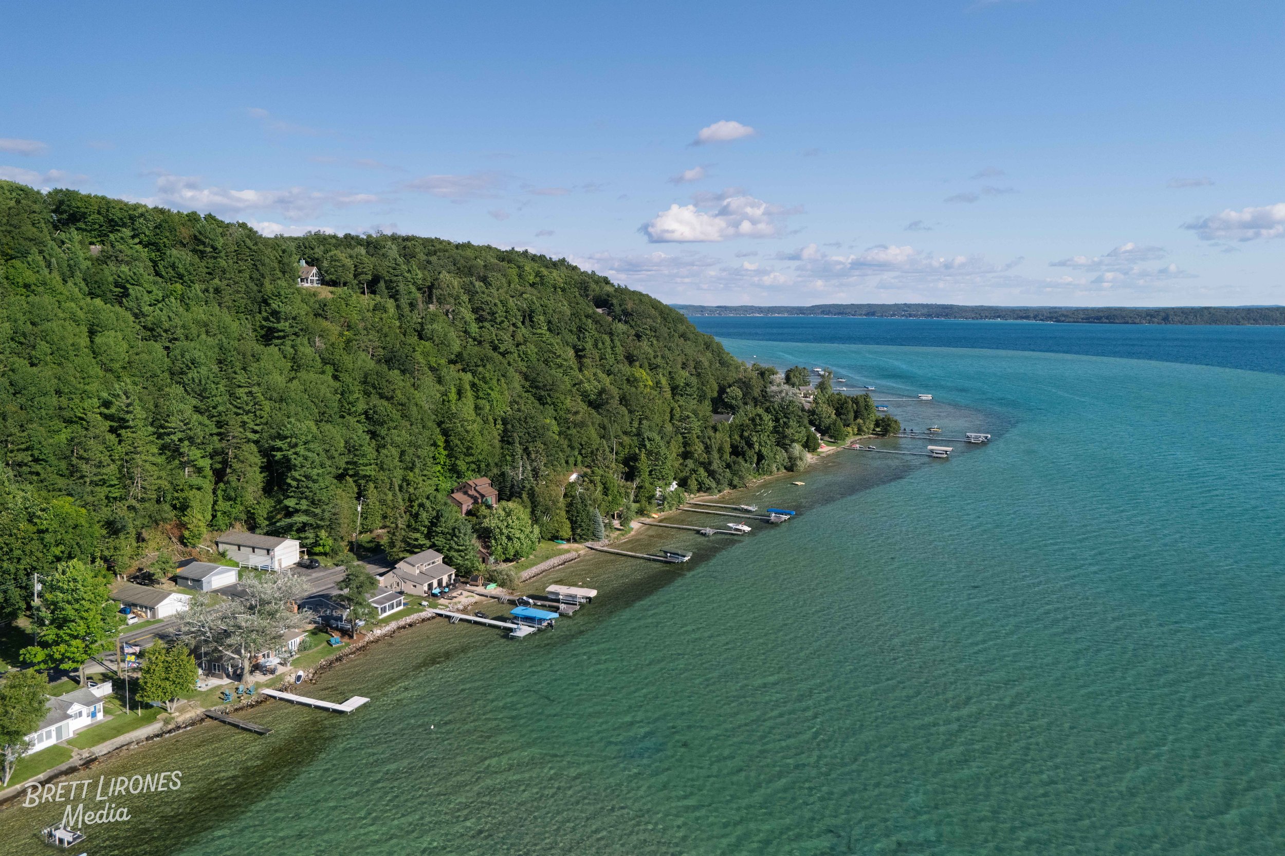 Aerial view of a lakeshore with houses and docks along a green forested shoreline, extending into calm blue-green water under a partly cloudy sky.