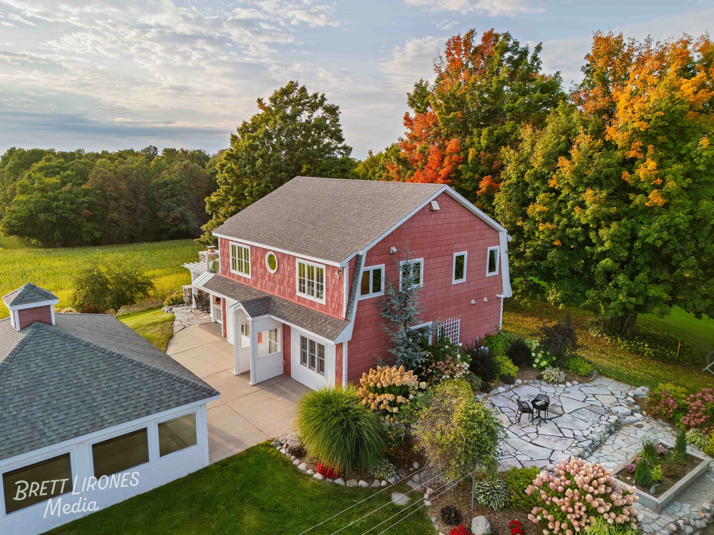A pink house with a gray roof surrounded by green trees and a landscaped garden with flowers and a patio with outdoor furniture.