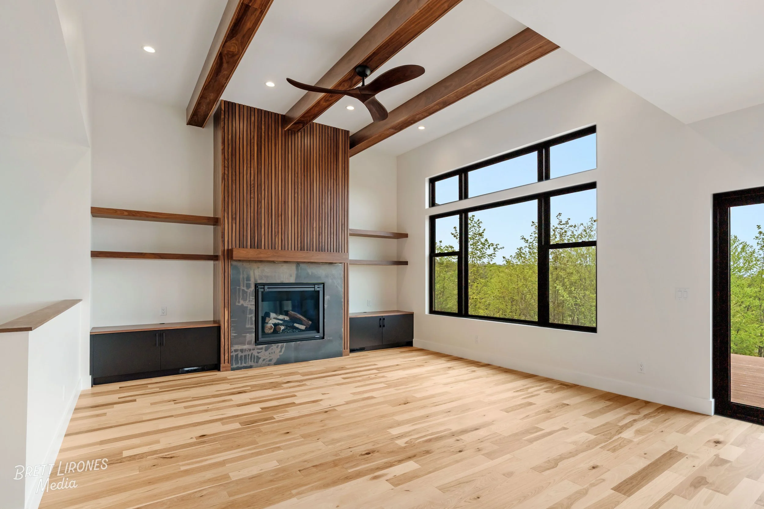 Empty living room with large black-framed window, a modern fireplace with a wooden surround, built-in shelving, exposed wooden beams, and light hardwood floors.