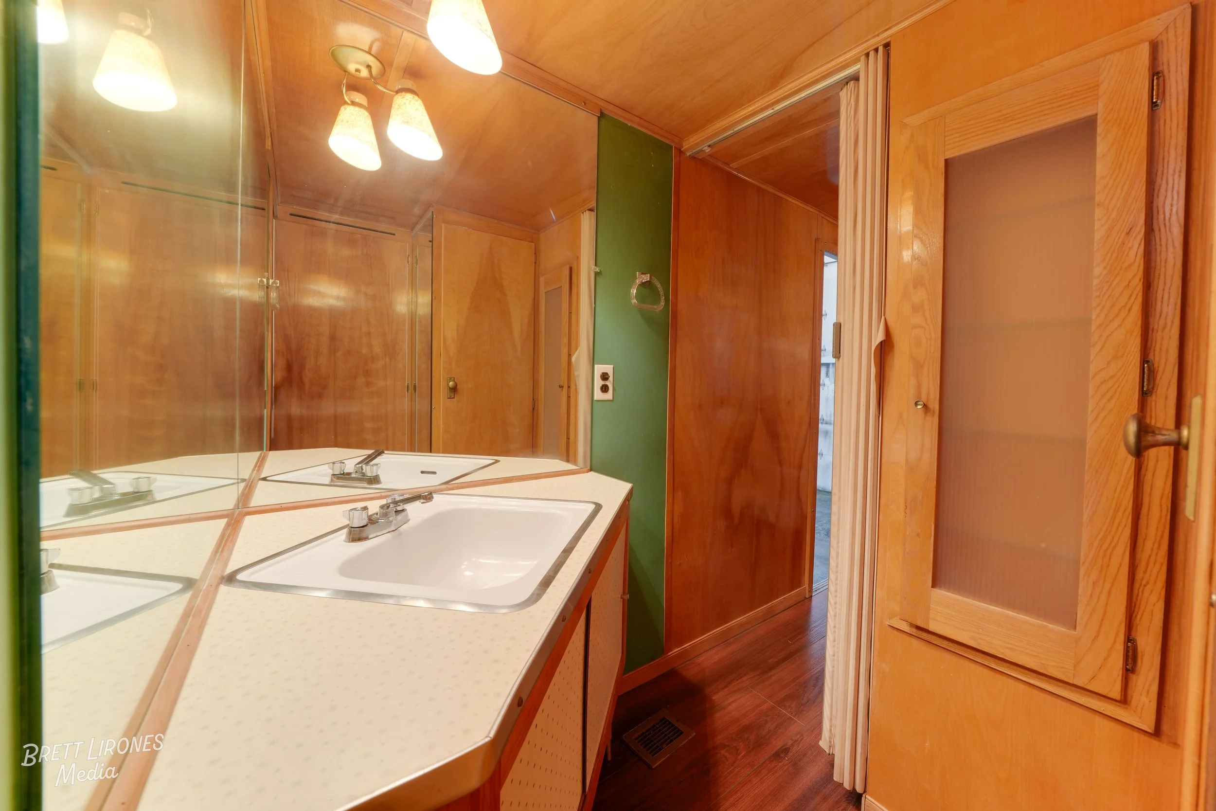 A bathroom with wood-paneled walls and a mirror above a white countertop with a sink. There is a light fixture with three lamps above the mirror, and a door with a frosted glass panel on the right side.