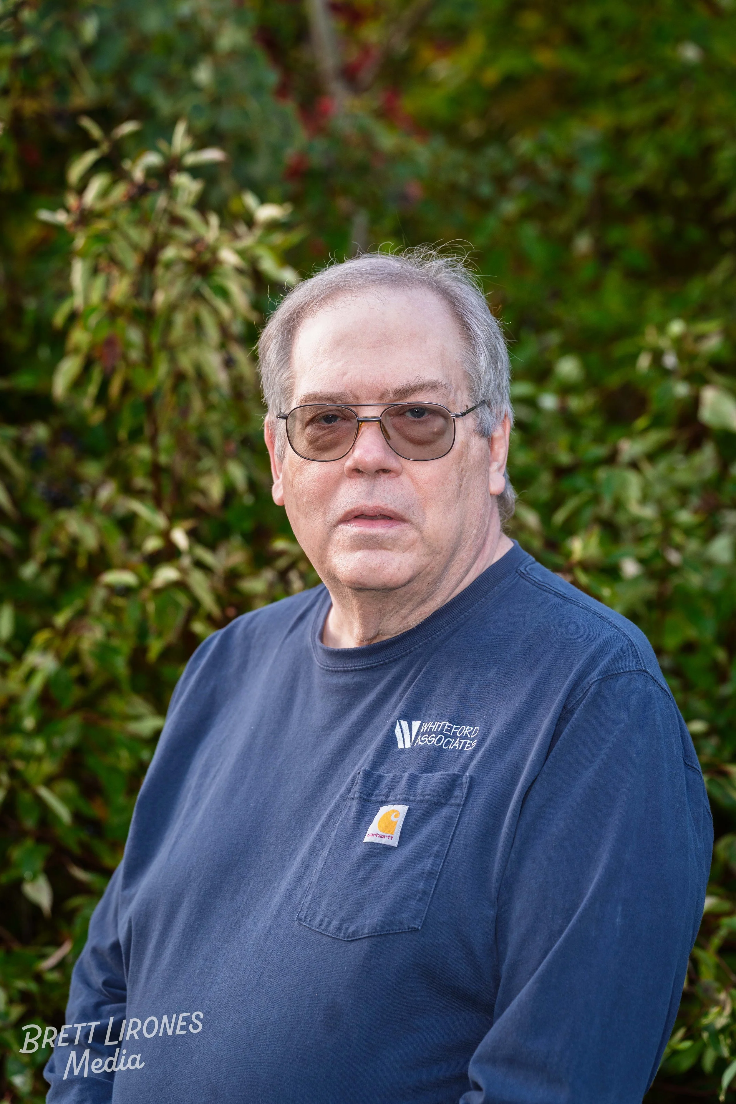 Older man with glasses wearing a navy blue Carhartt shirt and standing outdoors in front of bushes.