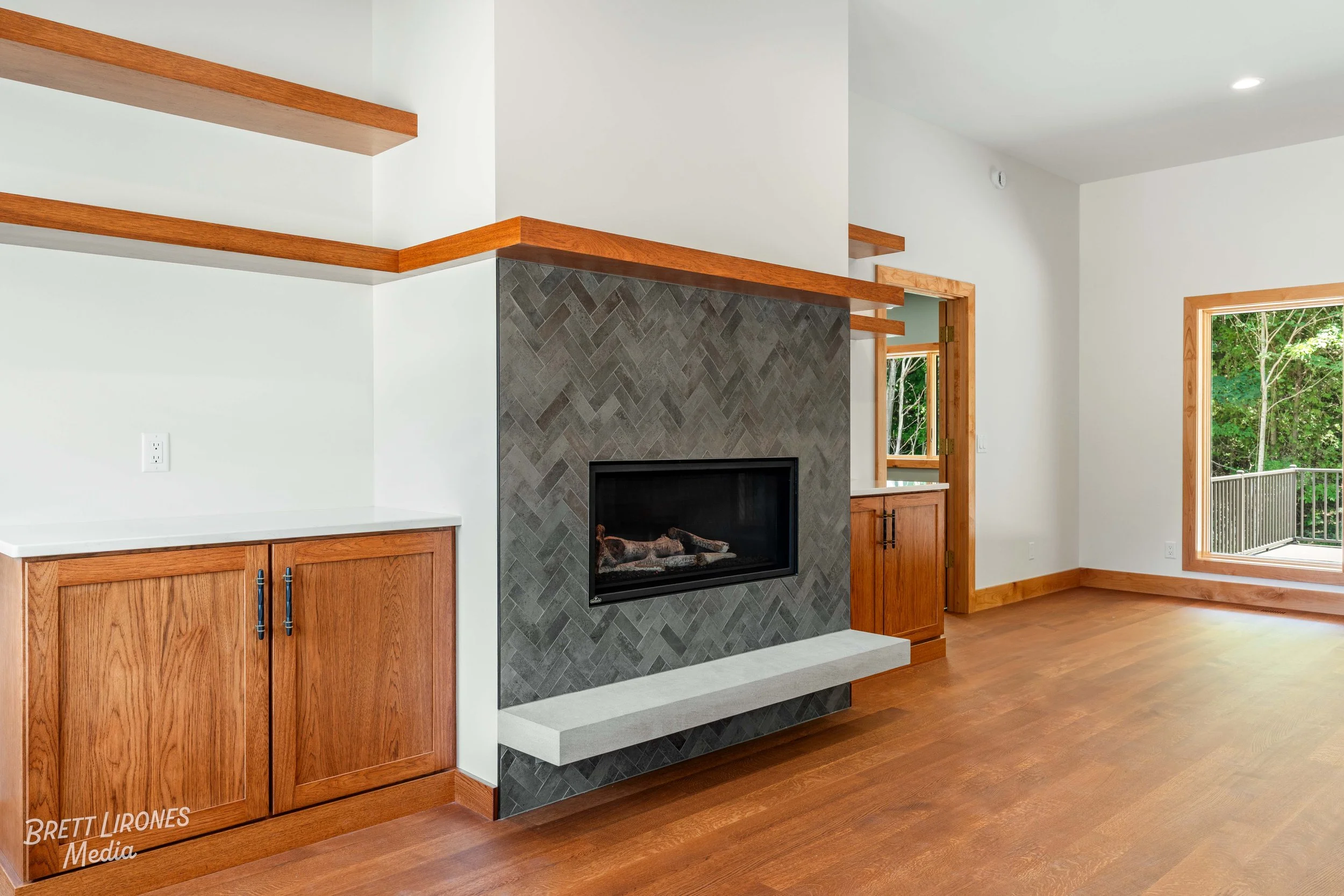 Interior of a living room featuring a modern black and gray tiled fireplace with a white stone hearth, surrounded by wooden cabinetry and trim, and a large window showing a view of trees outside.