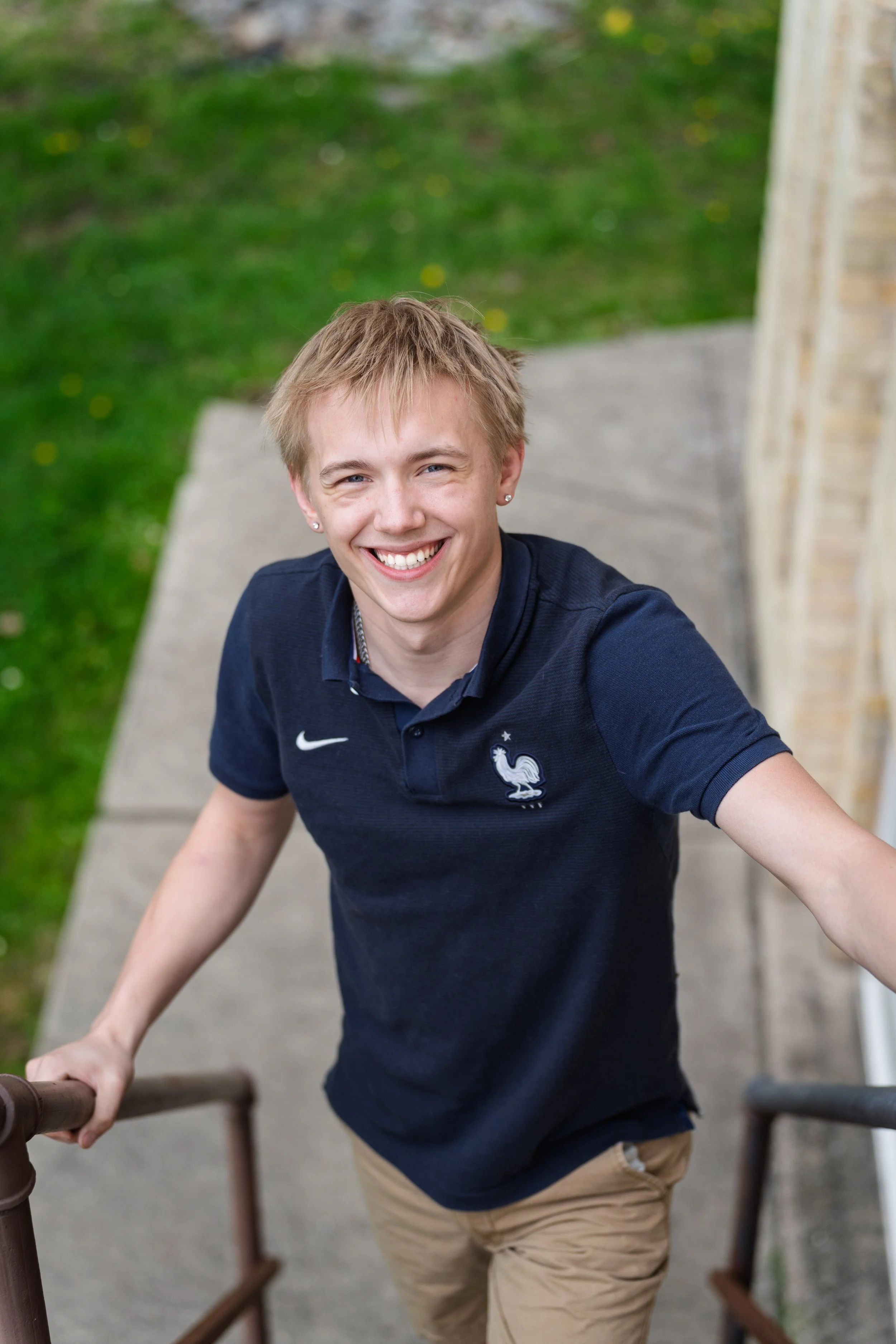 A young person with short blond hair, smiling and looking up at the camera, standing on outdoor stairs with a grassy area behind them, wearing a navy blue polo shirt with a rooster logo and beige pants.