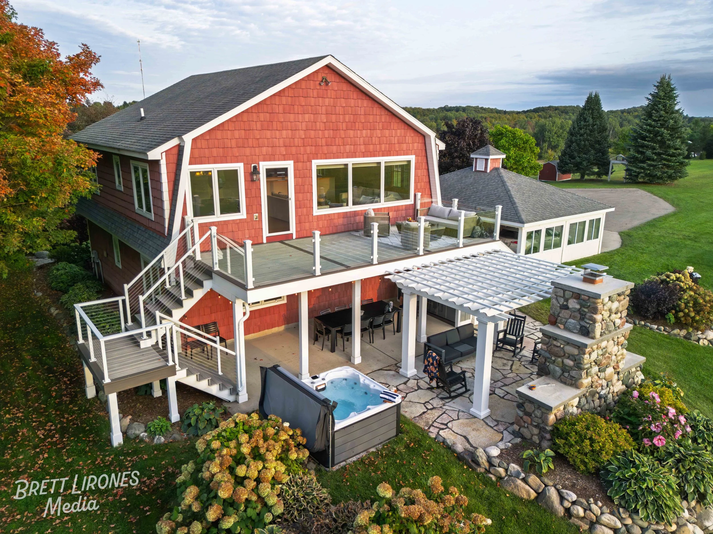 A two-story red house with a large outdoor deck and patio area, surrounded by green lawns and trees, with outdoor furniture, a hot tub, and a stone fireplace.