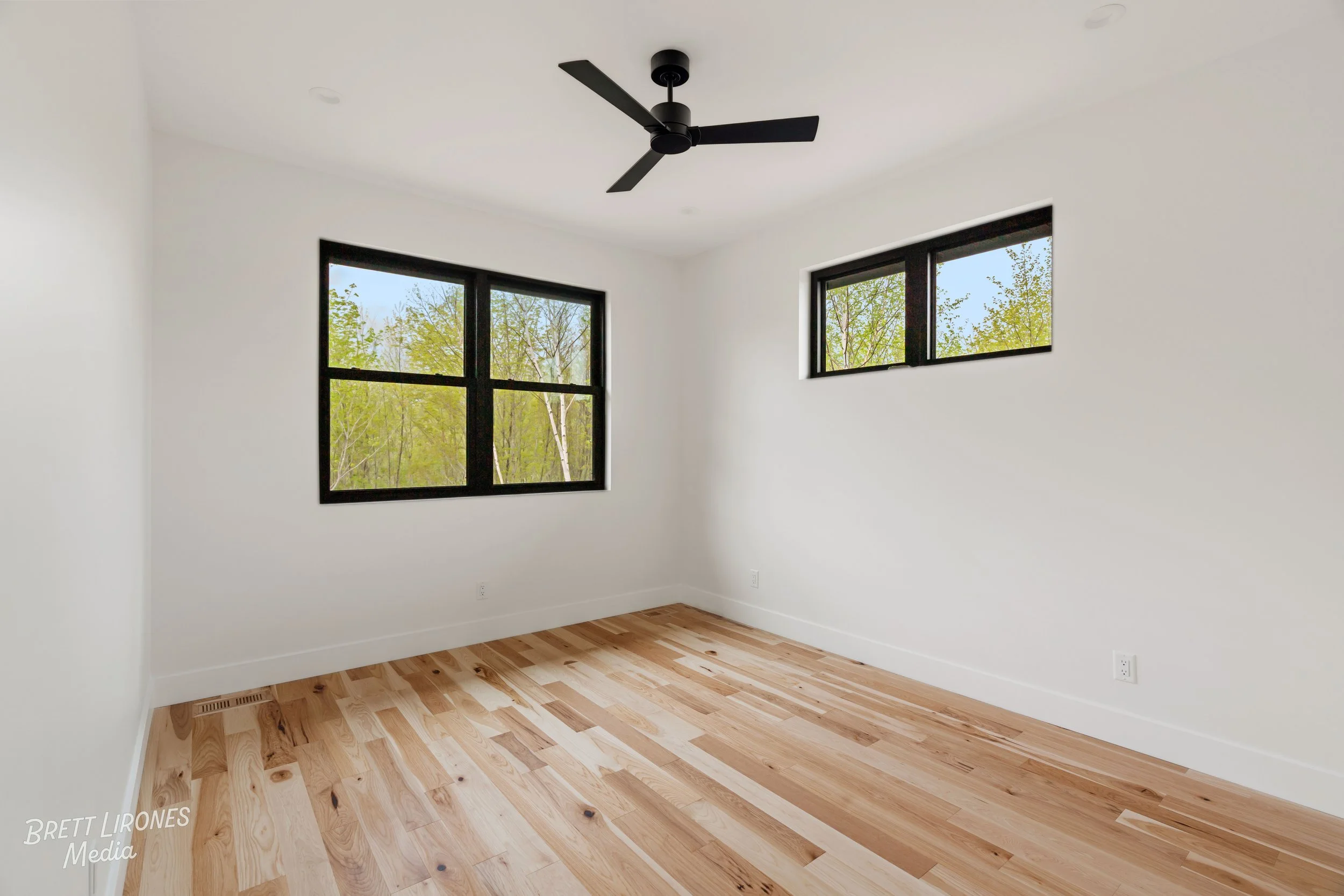 Empty room with white walls, hardwood floors, two black-framed windows showing green trees outside, ceiling fan, and electrical outlets.