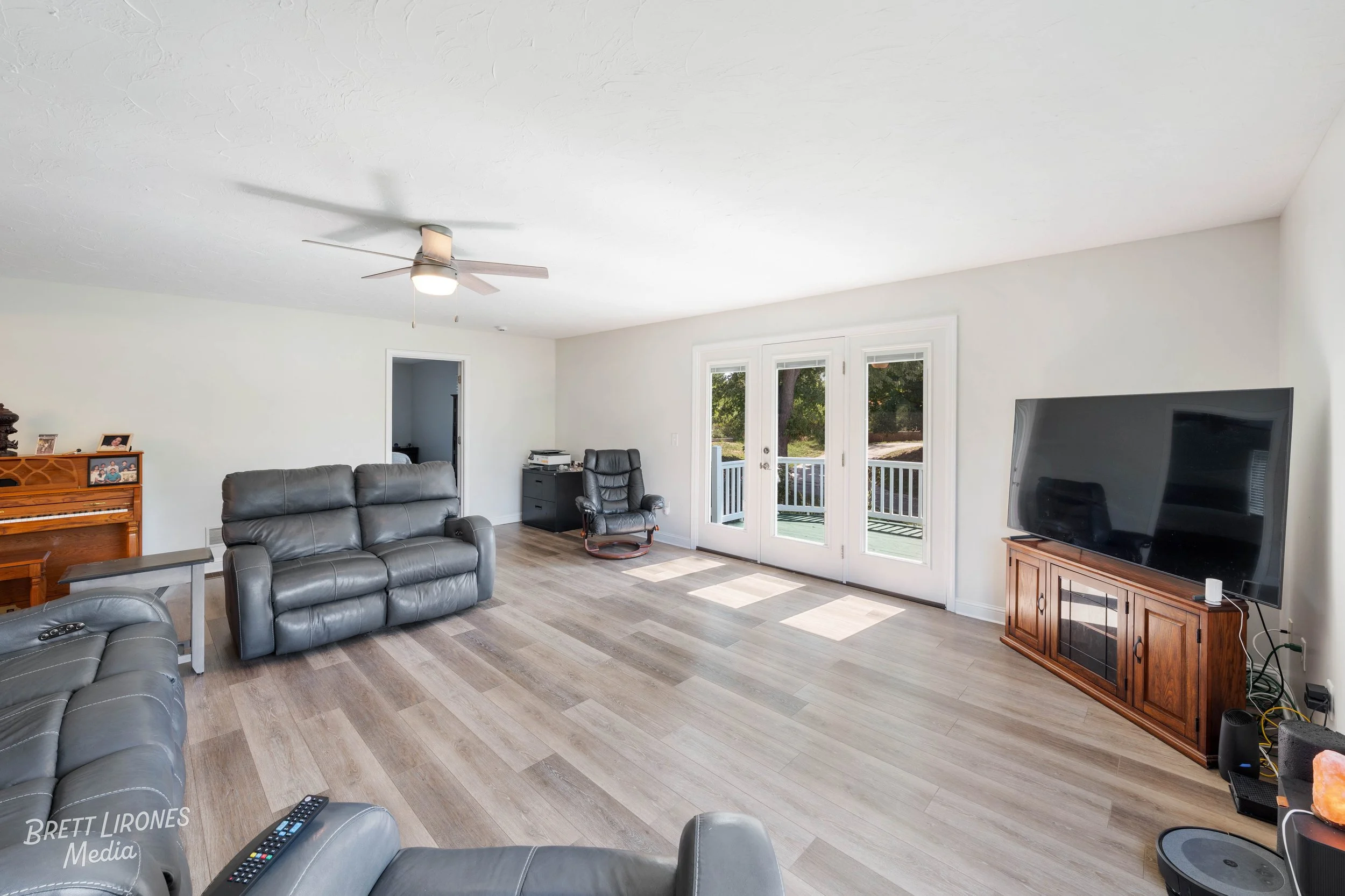 Living room with gray leather couches, wooden TV stand, large flat-screen TV, French doors leading outside, ceiling fan, hardwood floors, and some furniture near the walls.