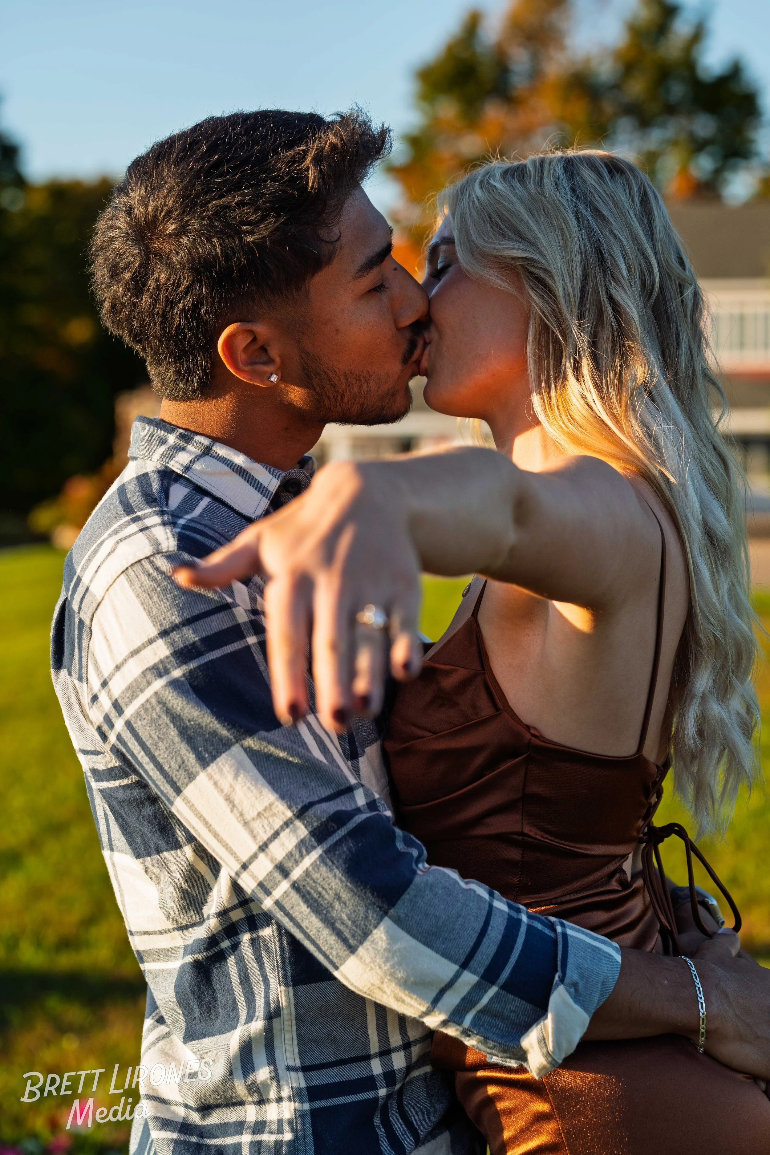 A young couple kissing outdoors during sunset, with the woman sitting on the man's lap, arms around him, in a park with trees and a house in the background.
