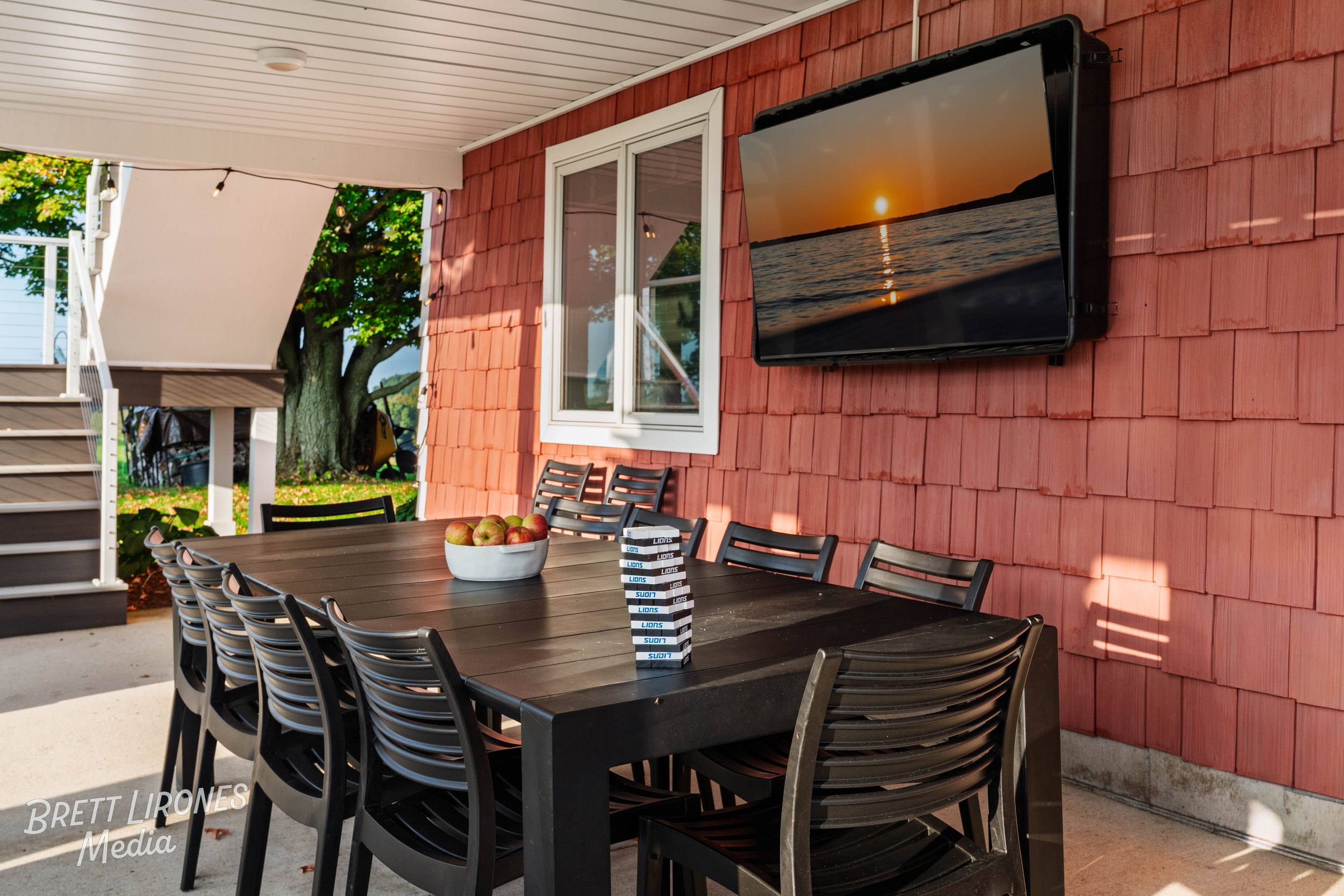 Outdoor dining area on a patio with a black table, black chairs, a bowl of apples, and a Jenga game, adjacent to a red wooden house wall with a mounted TV showing a sunset over water, and a window beside the TV.