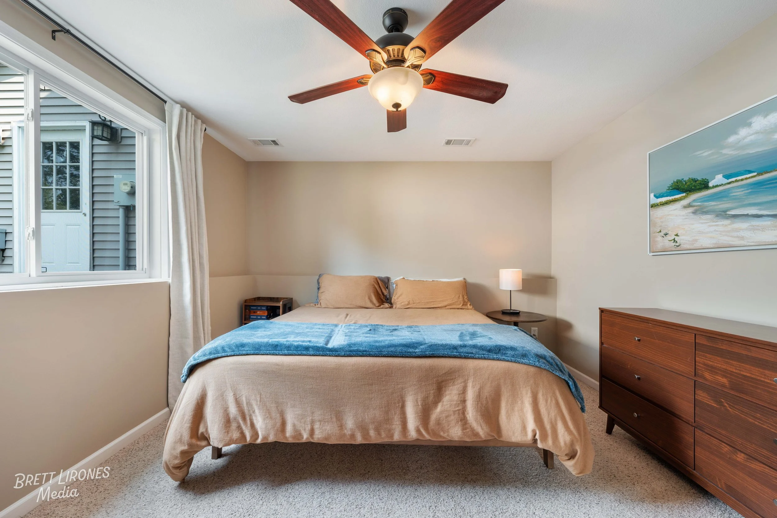 A bedroom with a bed covered in beige bedding, two beige pillows, a blue throw blanket at the foot of the bed, a wooden nightstand with a small lamp, a wooden dresser, a window with white curtains, and a painting of a beach scene on the wall, with a 