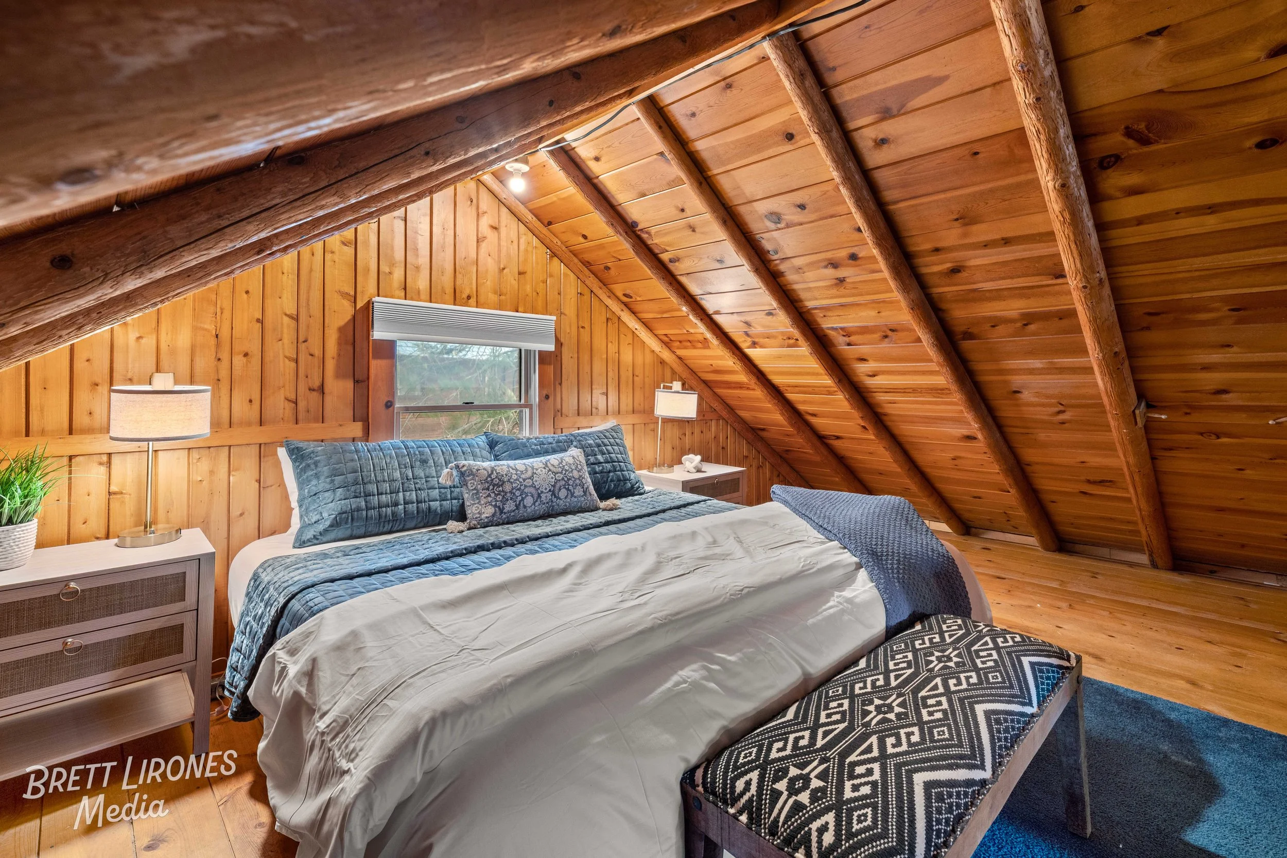 A rustic bedroom with wooden walls and sloped ceiling, featuring a large bed with blue and white bedding, two nightstands with lamps, a window above the bed, a patterned bench at the foot of the bed, and a small plant on one nightstand.