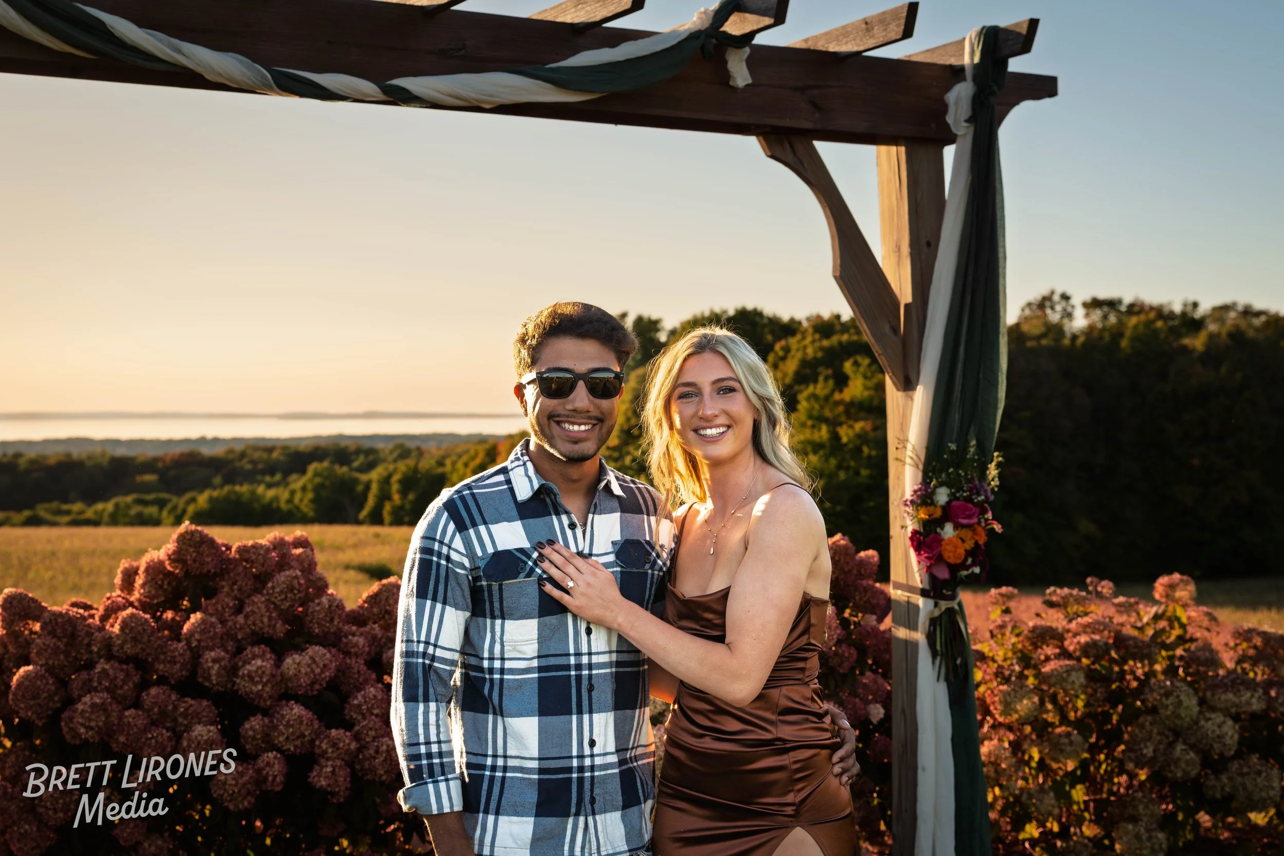 A smiling couple stands under a wooden wedding arch decorated with flowers during sunset in a field of hydrangeas with a distant tree line.