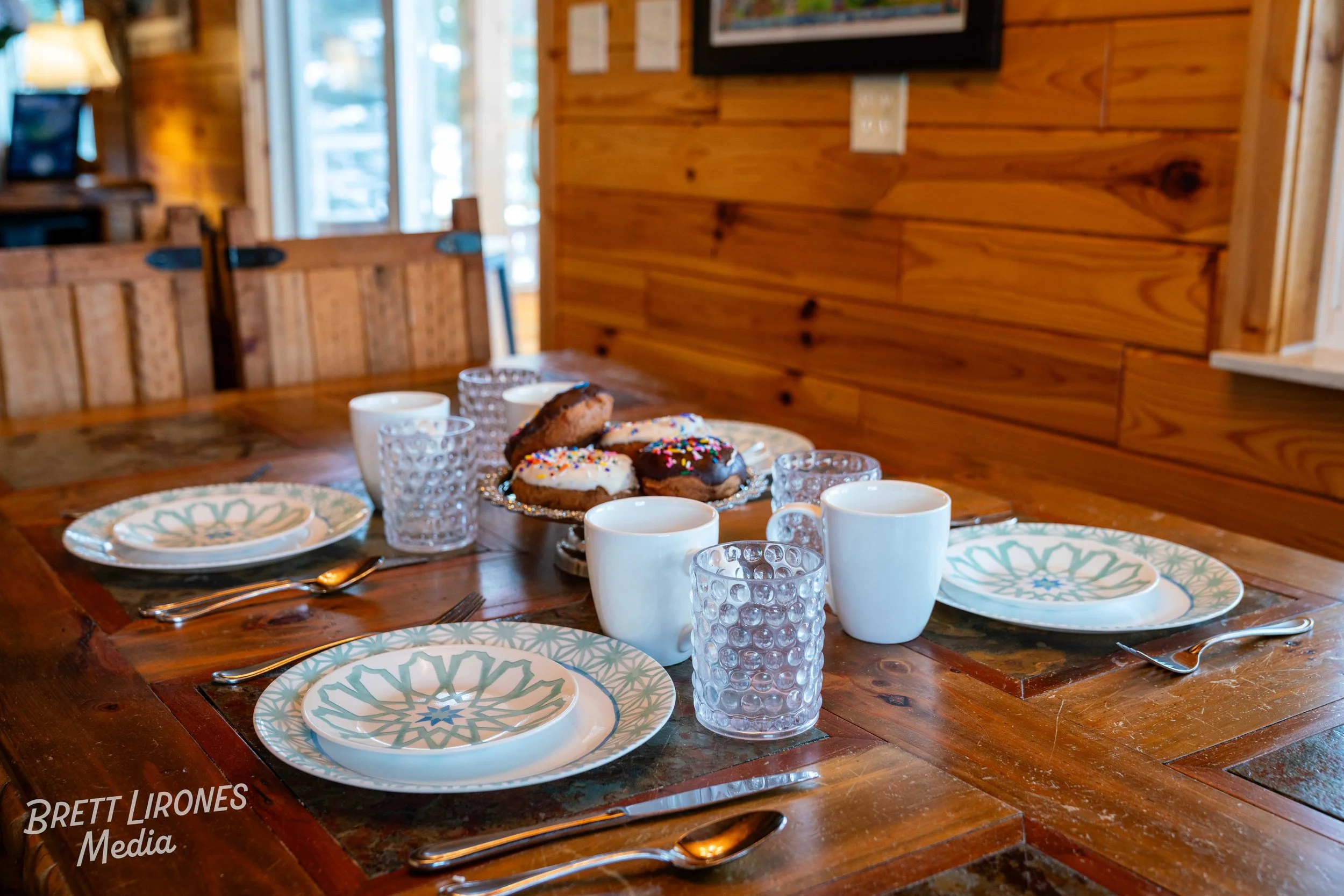 A wooden dining table set with plates, glasses, mugs, and a plate of assorted cookies with sprinkles, in a cozy cabin-style room.