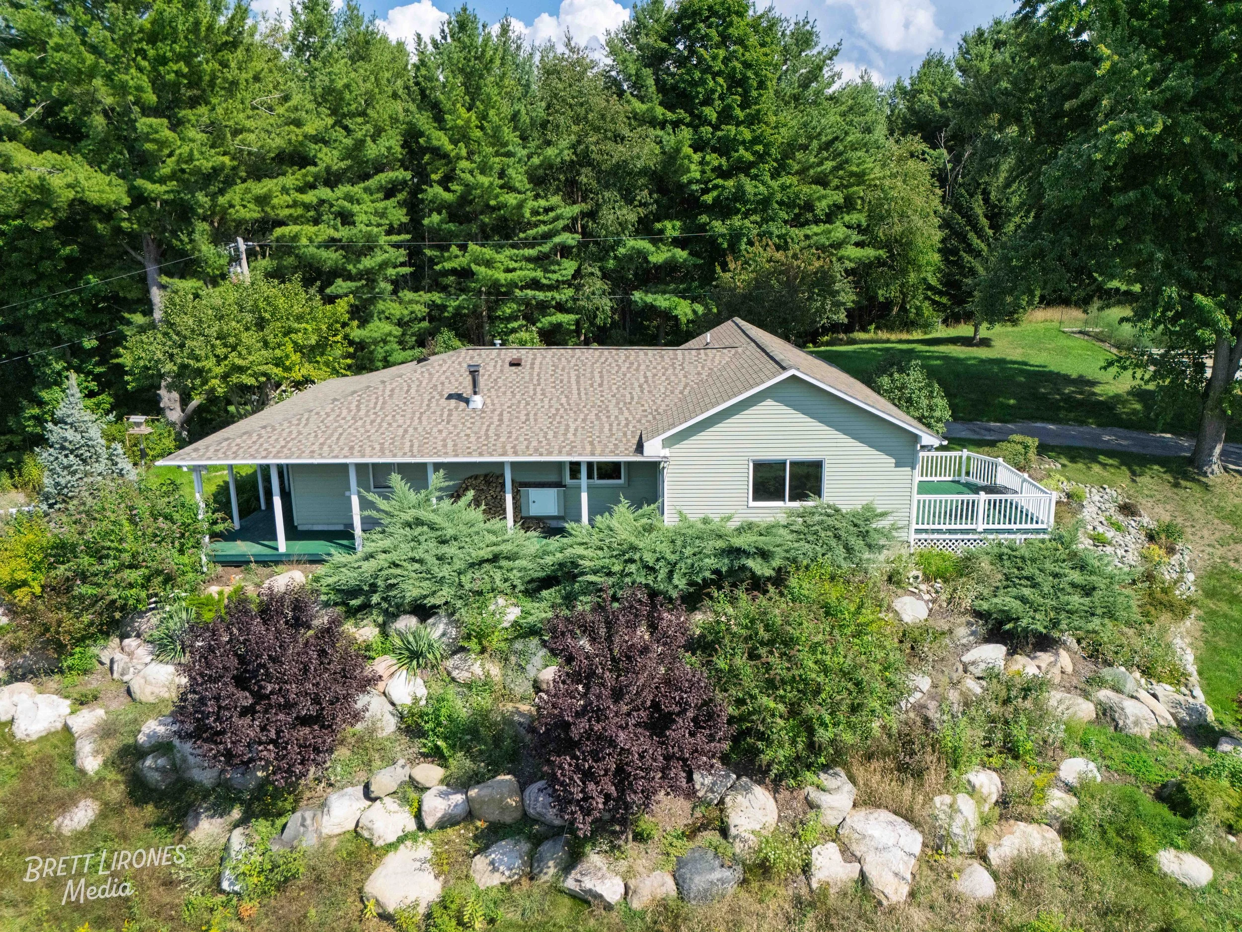 A house with a beige roof and light green siding, surrounded by trees and bushes, with a landscaped rock garden in the foreground.