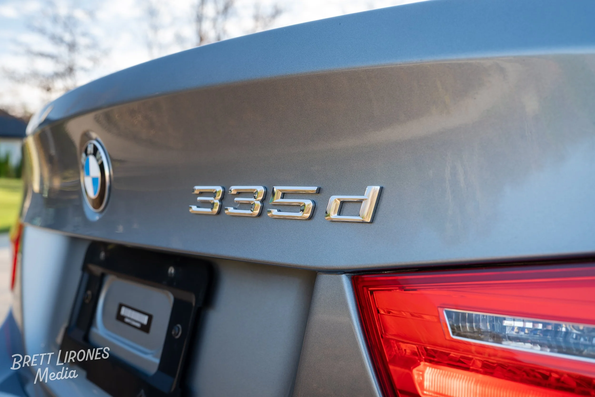 Close-up of the rear of a silver BMW 335d, showing the BMW emblem and model badge, with a red taillight in the bottom right corner.