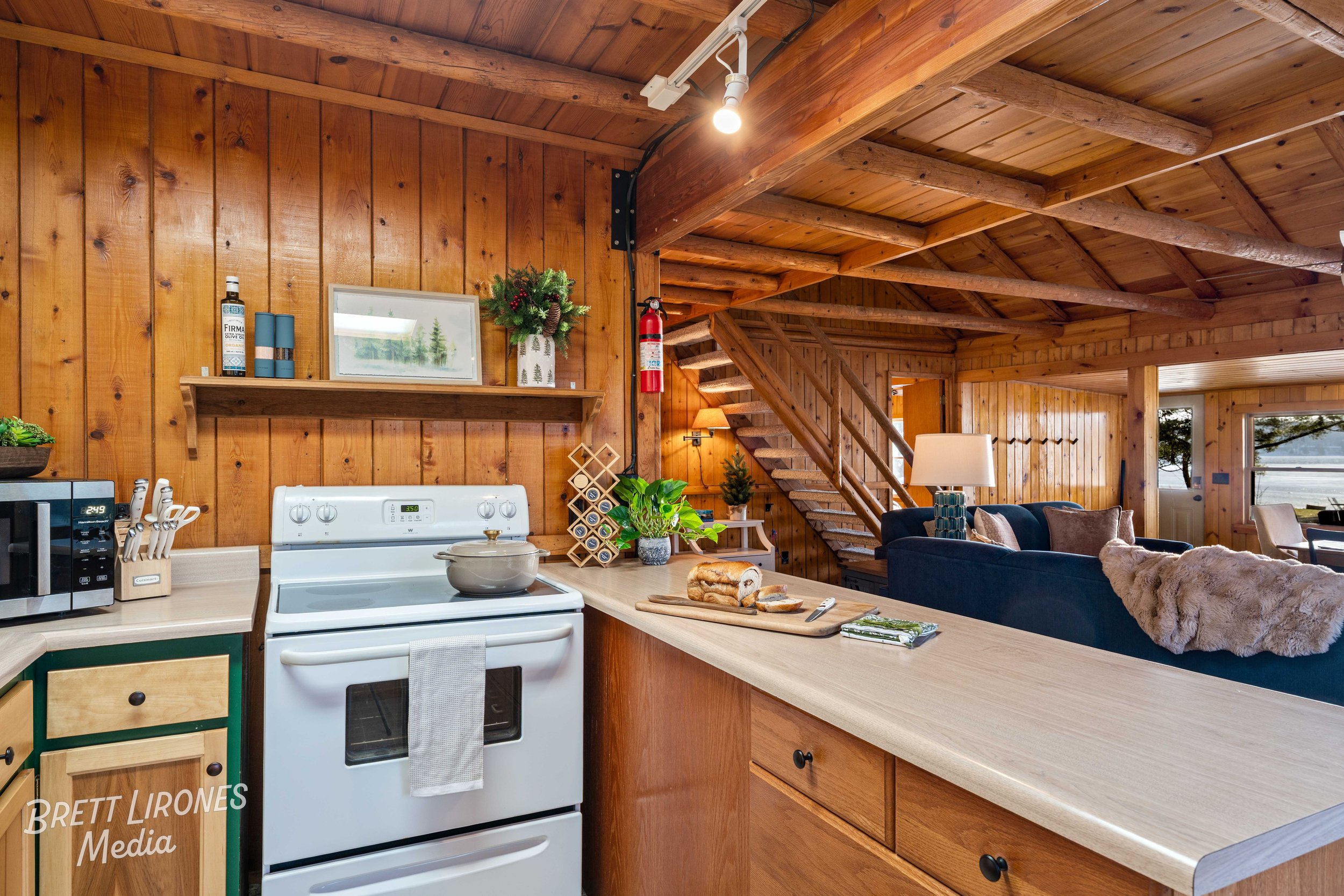 Interior of a cozy cabin kitchen with wooden walls and ceiling, featuring a white stove, microwave, cutting board with baked goods, and various decor items, with a living area and staircase in the background.