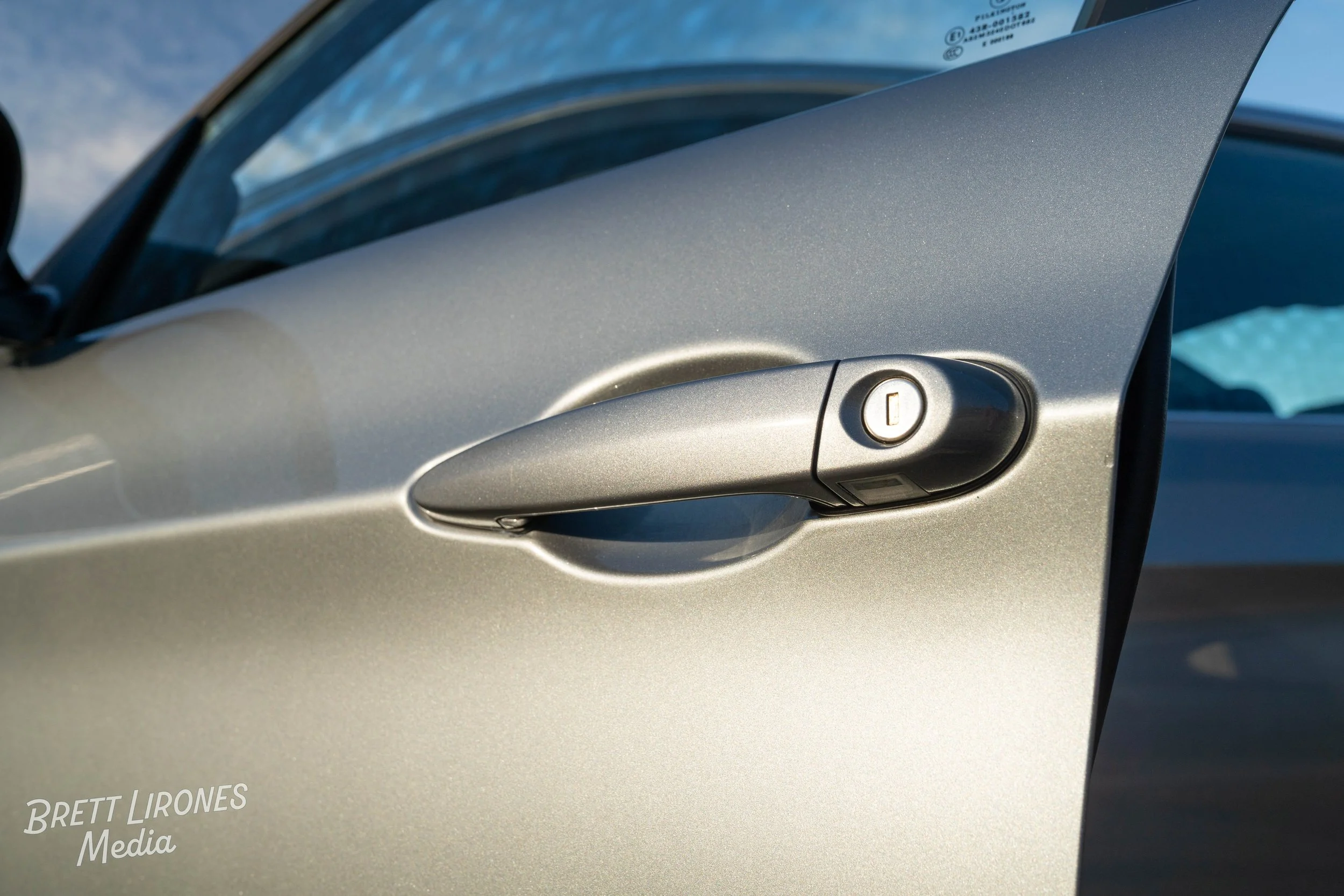 Close-up of a silver car door handle with a lock/unlock button, on a silver vehicle, with a partly visible windshield and sky in the background.
