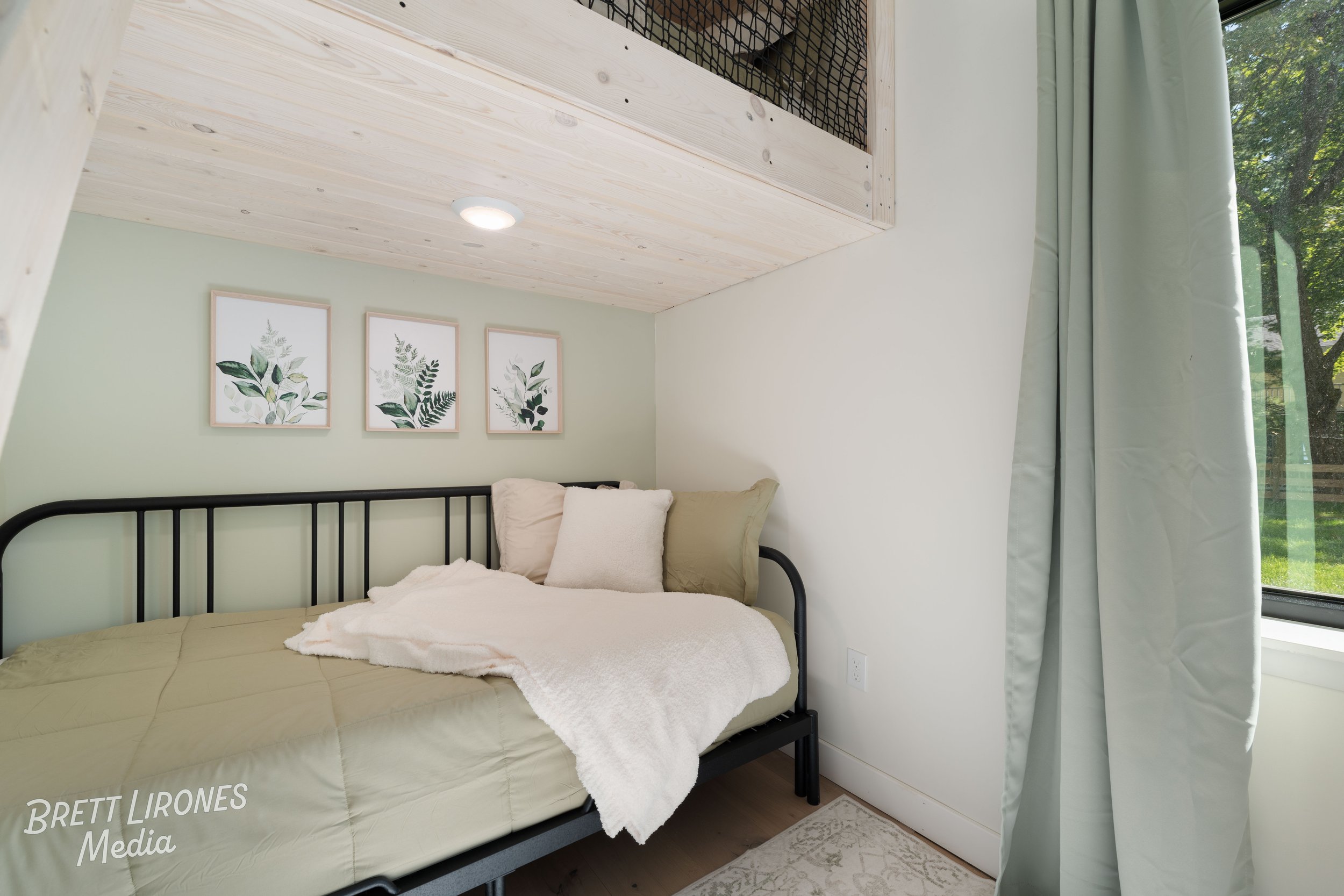 A bedroom with a metal bed frame, beige bedding, and multiple pillows against a green wall with three botanical art prints. A window with light-colored curtains shows a view of trees outside.