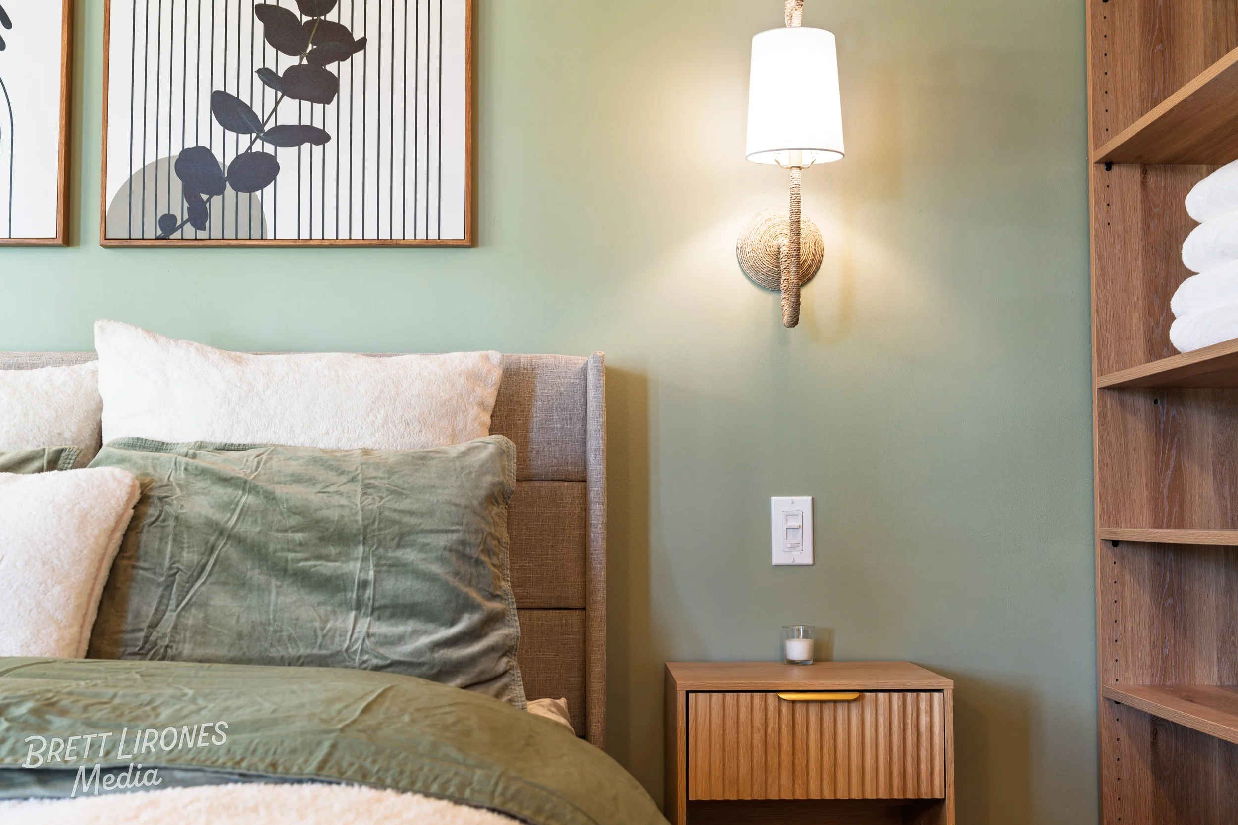 A bedroom with a bed featuring multiple pillows against a green wall. There is a wooden nightstand with a small candle, a wall-mounted light fixture with a white shade and rope detail, and two framed pieces of art with botanical designs. Part of a wo