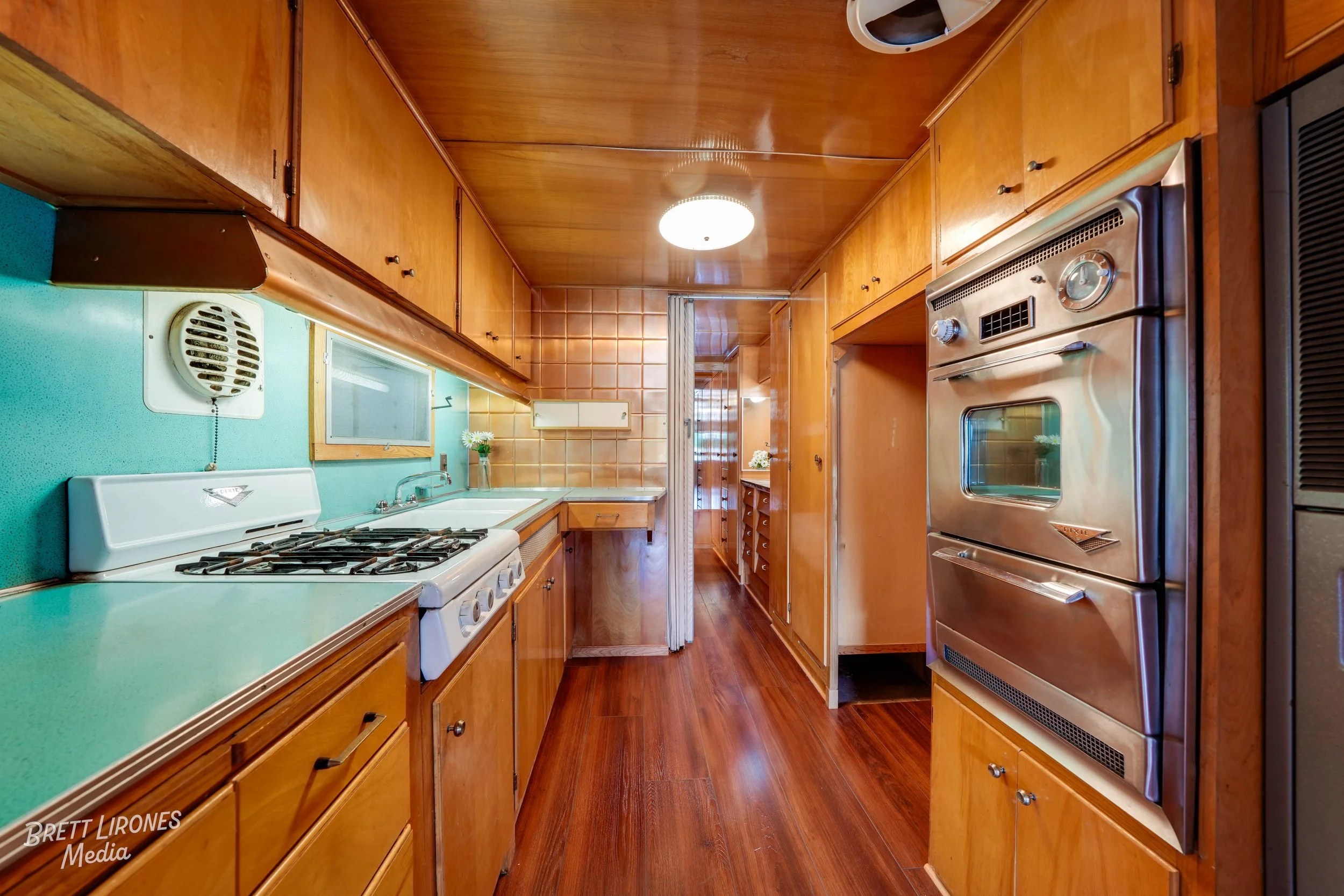Vintage kitchen with wooden cabinets, gas stove, green countertops, teal wall, window, and wood floor.