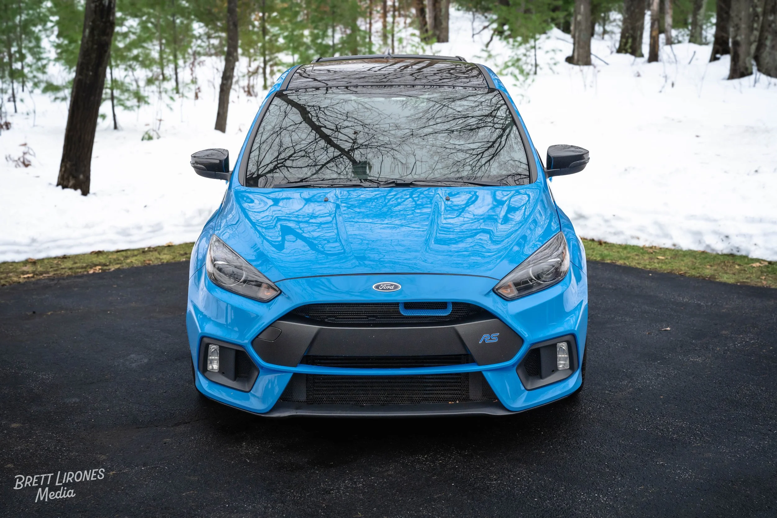 Front view of a blue Ford Focus RS parked on black asphalt with snow and trees in the background.