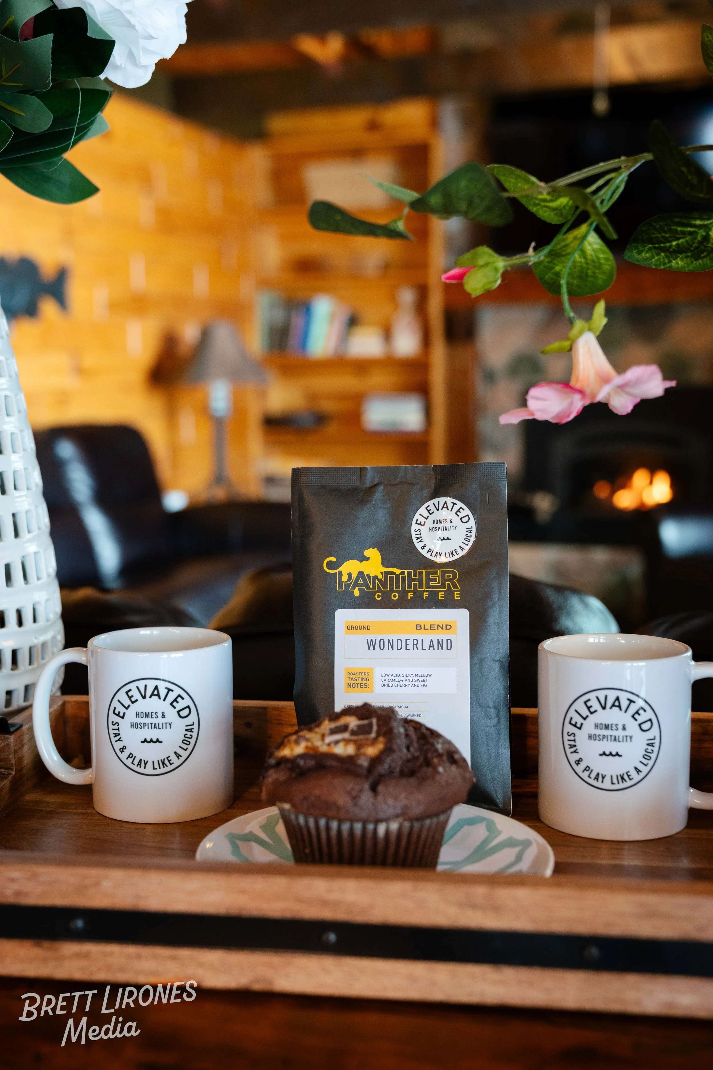 A tray with two white mugs marked 'Elevated Homes & Hospitality Stay & Play Like a Local,' a chocolate muffin topped with caramel and chocolate shavings on a plate, and a bag of Panther Coffee Wonderland ground coffee, set on a wooden surface in a co