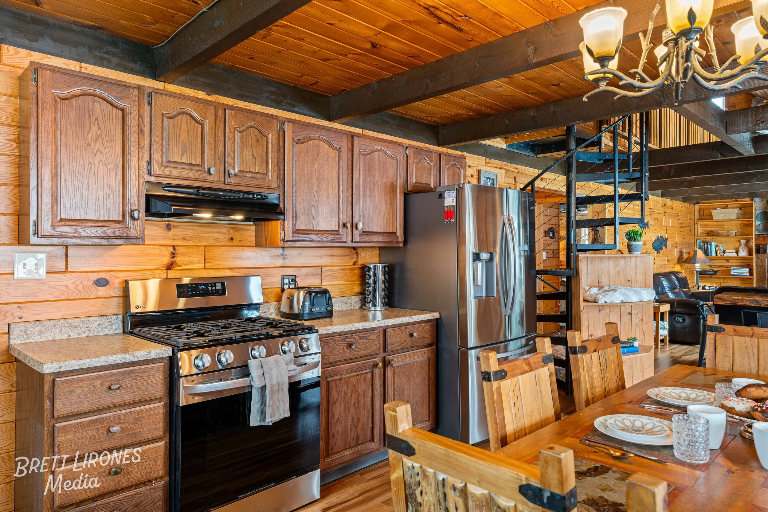 Kitchen with wooden cabinets, stainless steel refrigerator, stove, toaster, and spiral staircase in a rustic log cabin style.