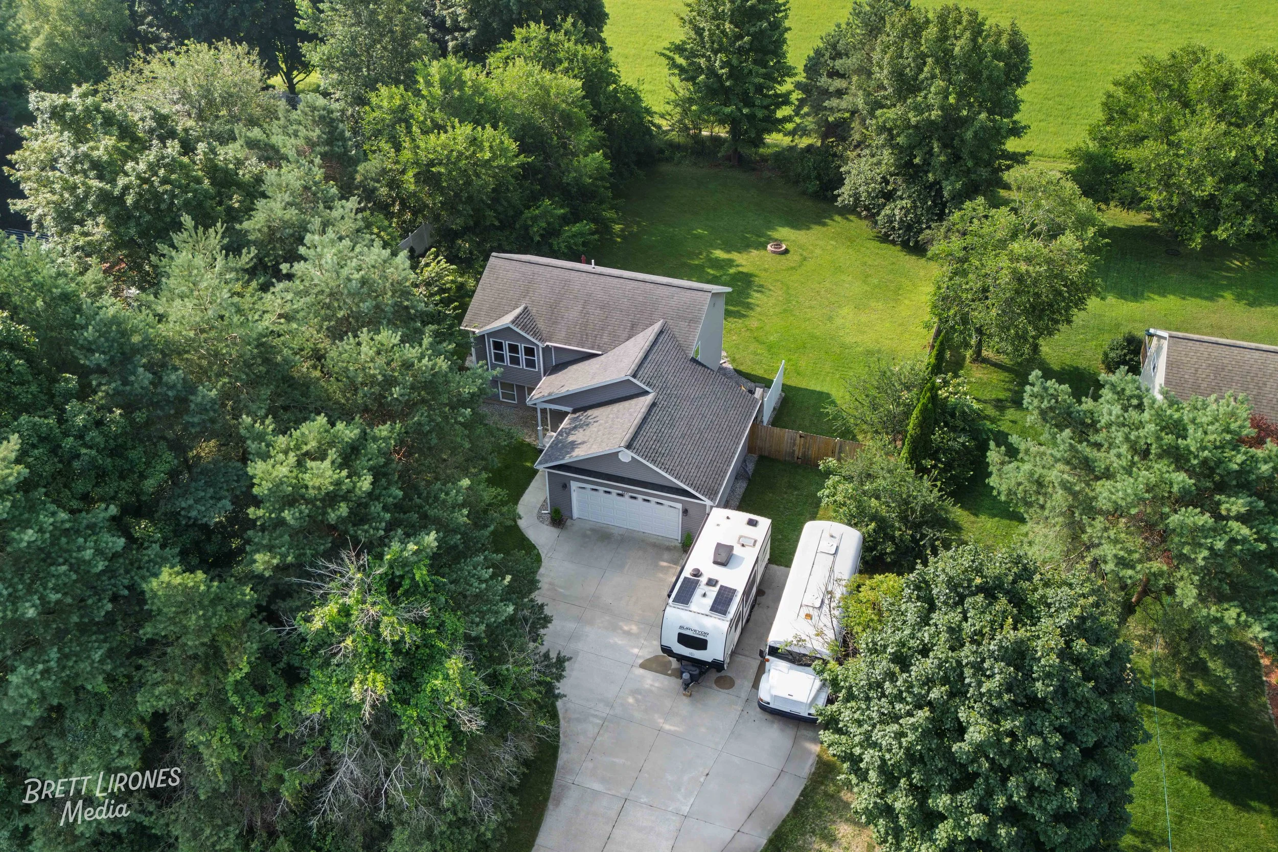 Aerial view of a house with a driveway, surrounded by trees and a grassy backyard with a fire pit.