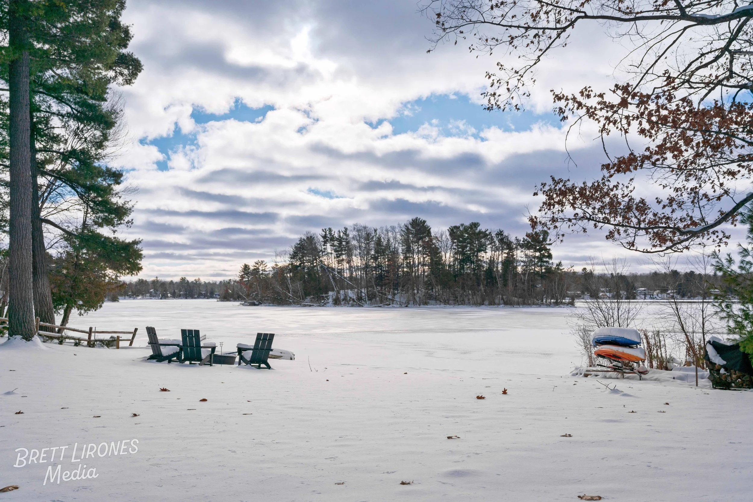Snow-covered lakeside with Adirondack chairs, boats on stands, and trees under a partly cloudy sky.