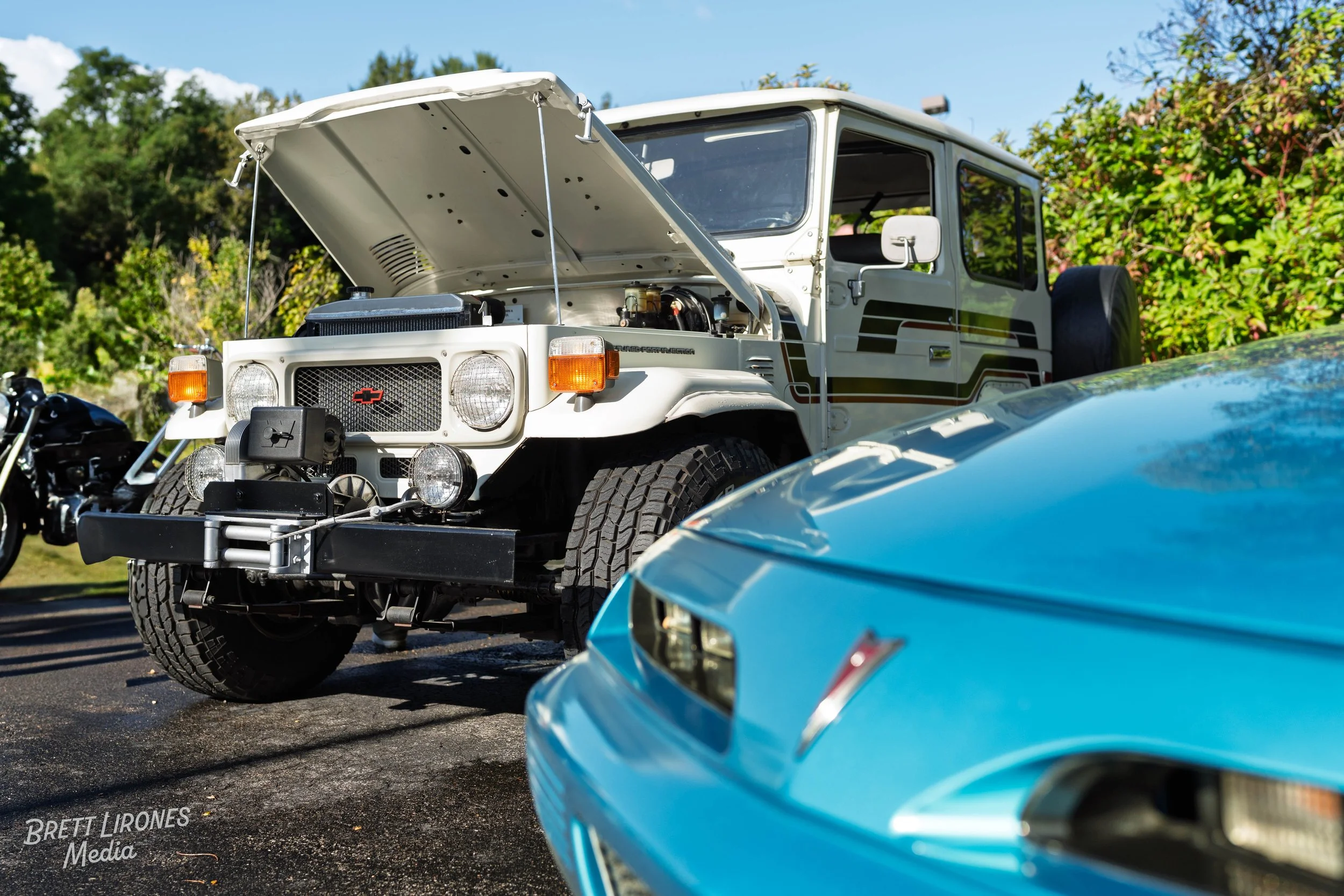 A white vintage off-road vehicle with its hood open, parked outdoors on pavement, alongside a blue sports car, with trees and a blue sky in the background.