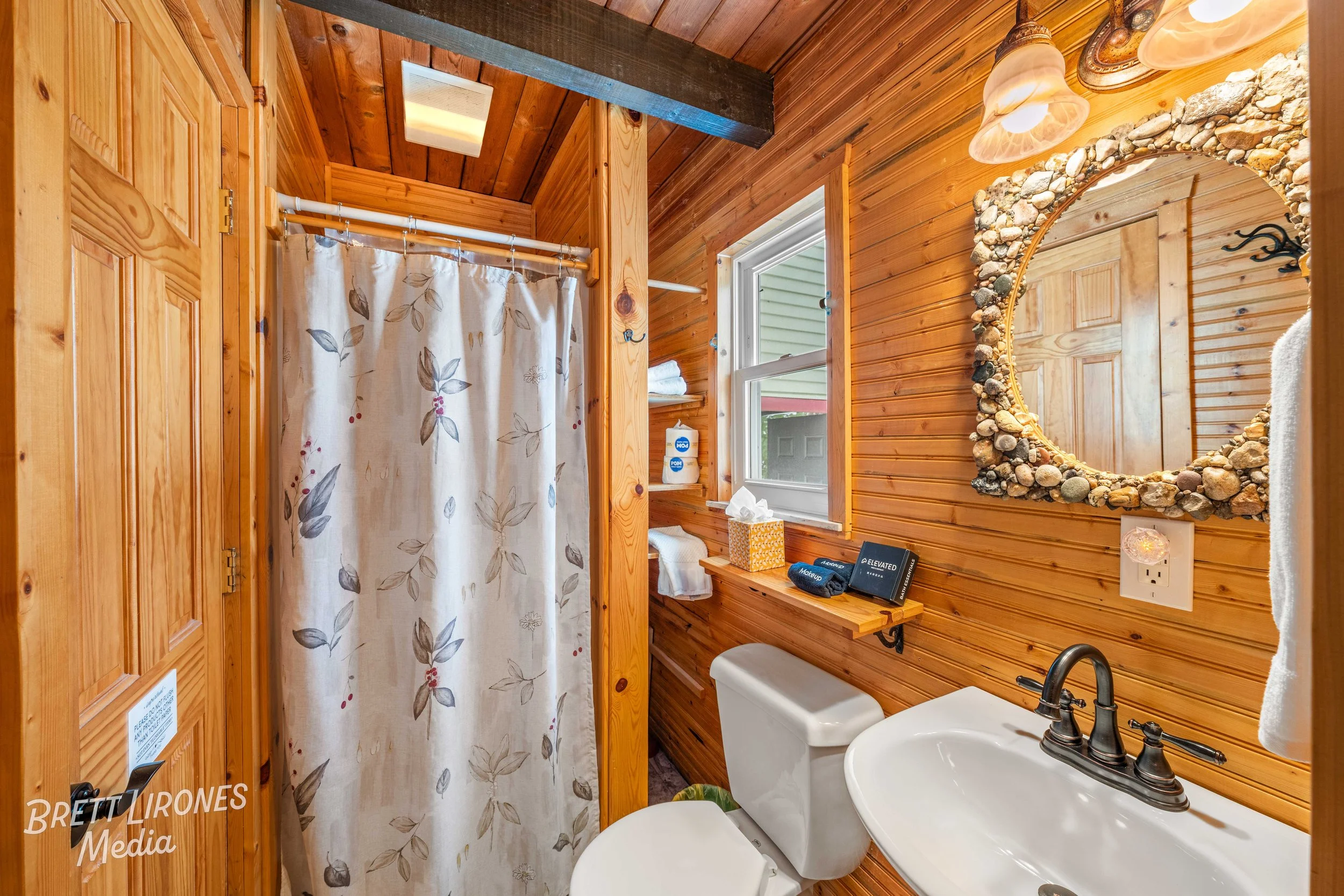 Wood-paneled bathroom with a shower with a floral curtain, a small window, a toilet, a sink with dark bronze fixtures, a mirror with a stone frame, and warm lighting.