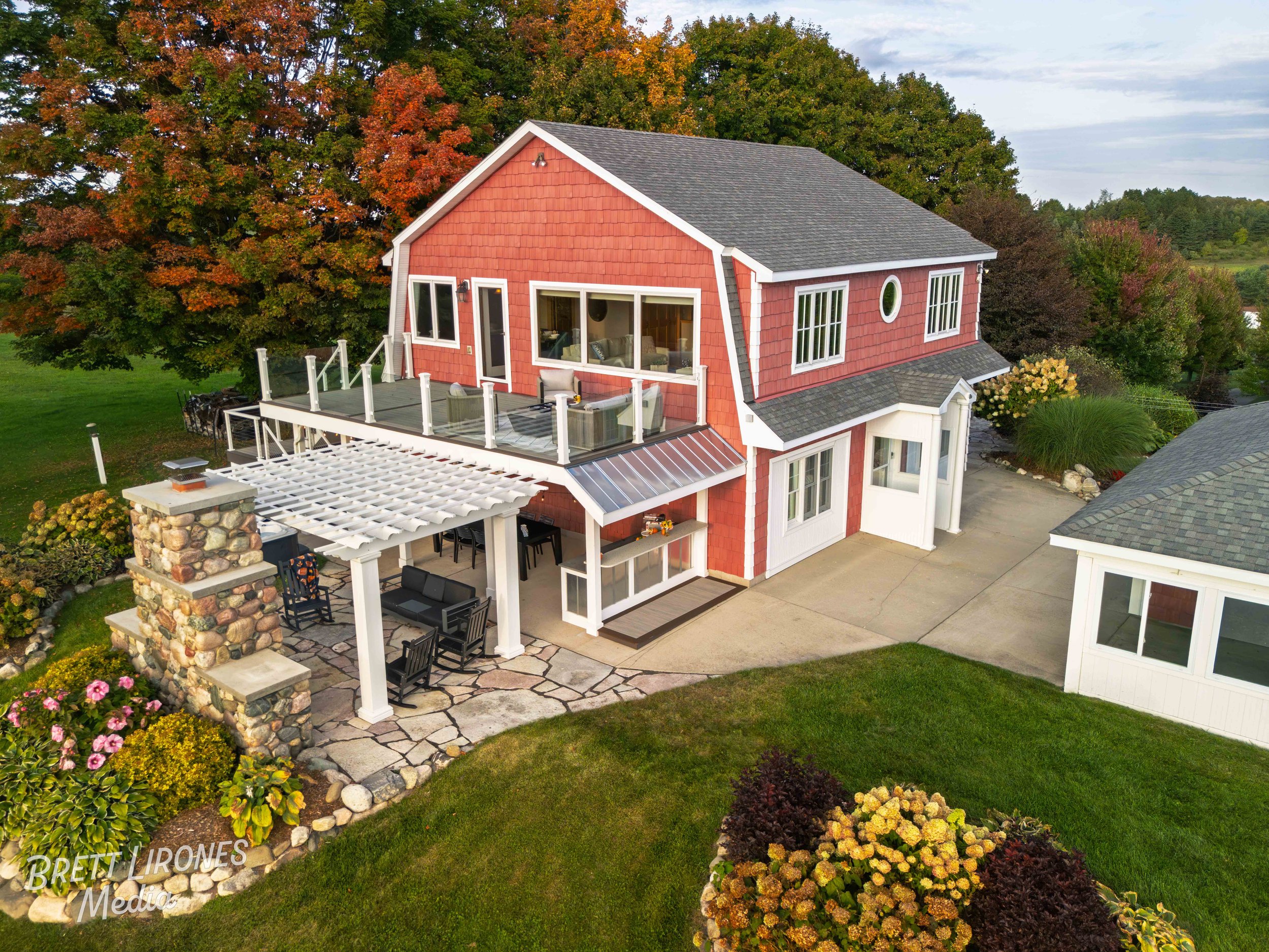 A two-story house with red siding, white trim, and a gray roof, featuring a second-floor deck with glass railing and outdoor seating, surrounded by a garden with colorful flowers and trees, with a paved patio and lawn.
