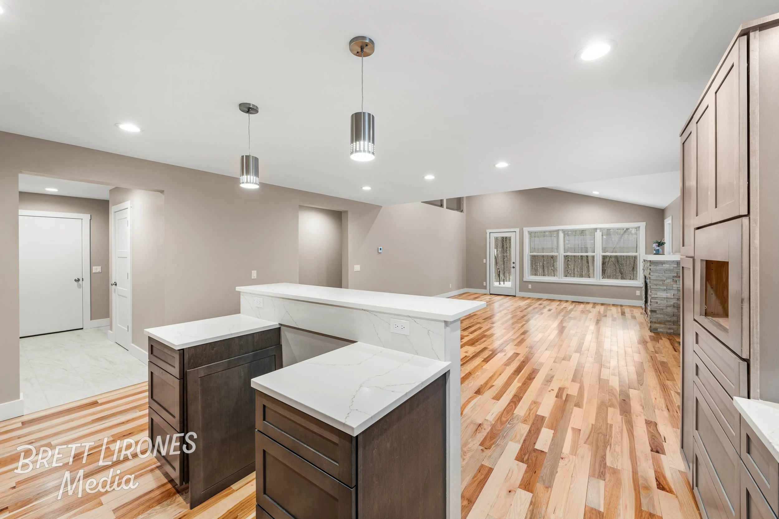 Modern open-concept living area with hardwood floors, large windows, and neutral-colored walls; kitchen island with white marble countertop and dark wood cabinets; twin pendant lights and recessed ceiling lights lighting the space.
