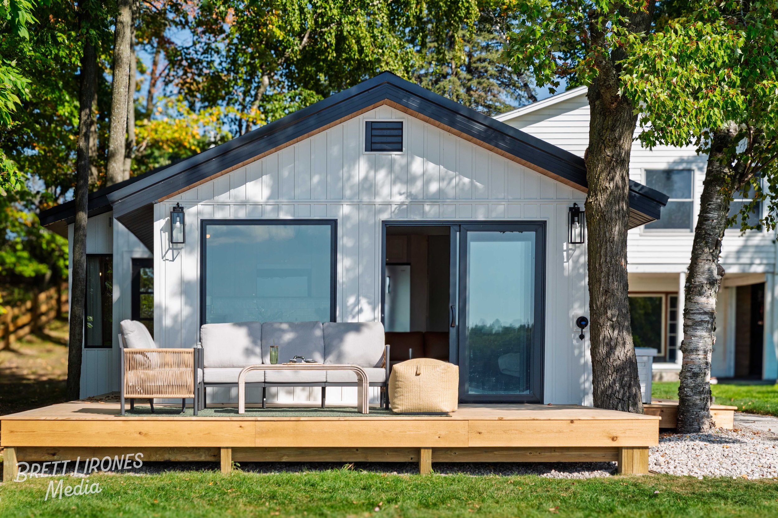 Small modern white house with black trim, large sliding glass door, and outdoor seating on wooden deck, surrounded by trees and green grass.