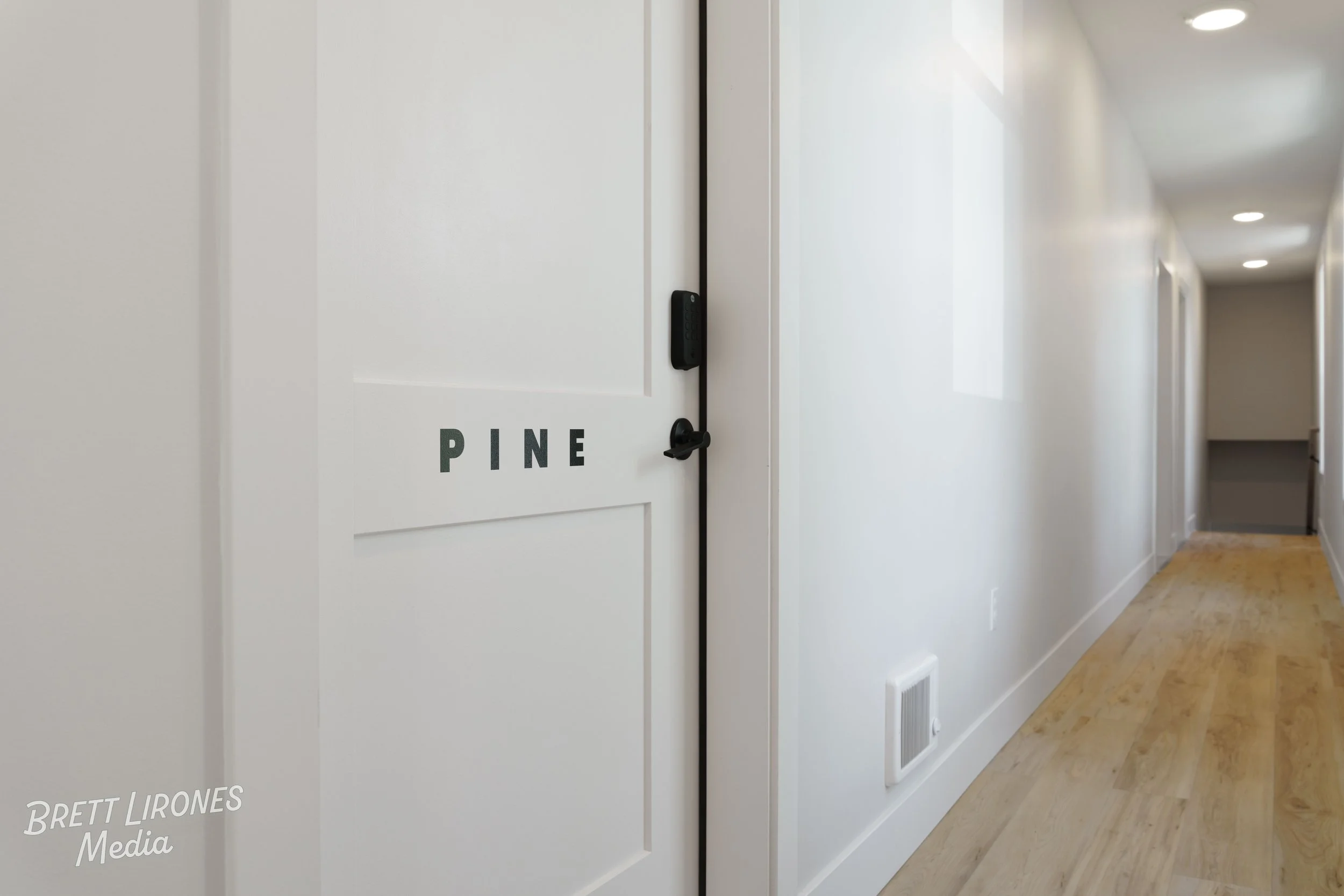 Interior view of a white door with the word 'PINE' on it, part of a hallway with white walls, wooden floor, and ceiling lights.