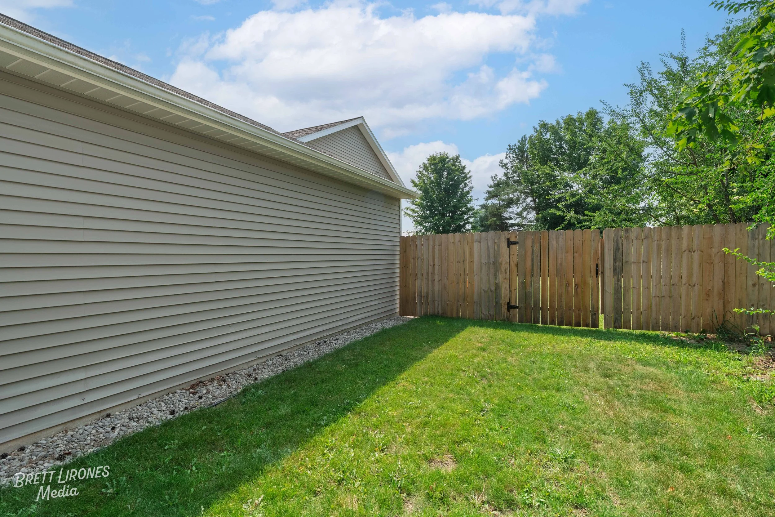Backyard with a grassy lawn, beige house siding on the left, and a wooden fence with a gate at the far end, surrounded by trees and blue sky with clouds.