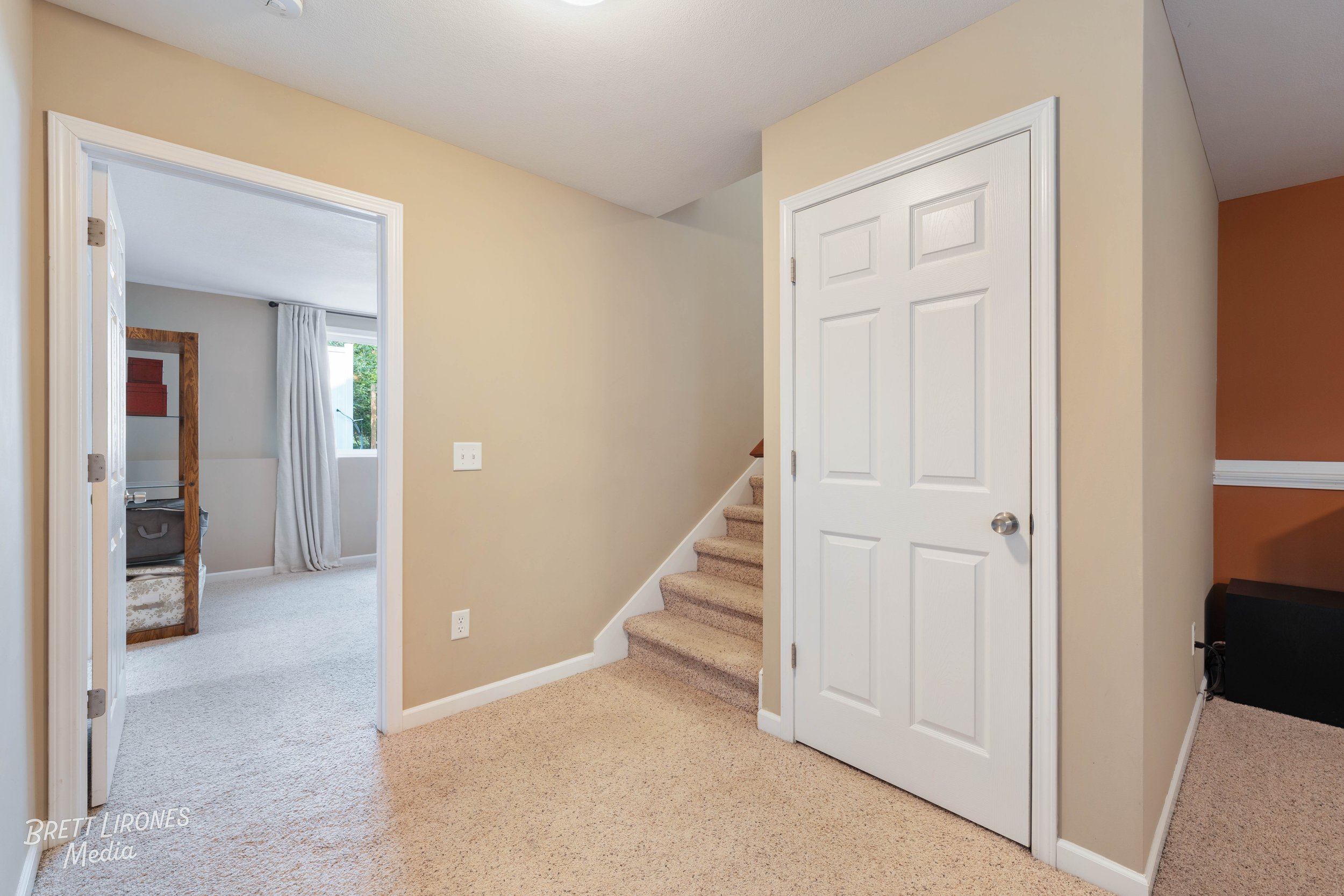 Interior view of a house showing a hallway with a staircase and two doorways leading to other rooms. The walls are painted in neutral tones, and the floors are covered with beige carpet.